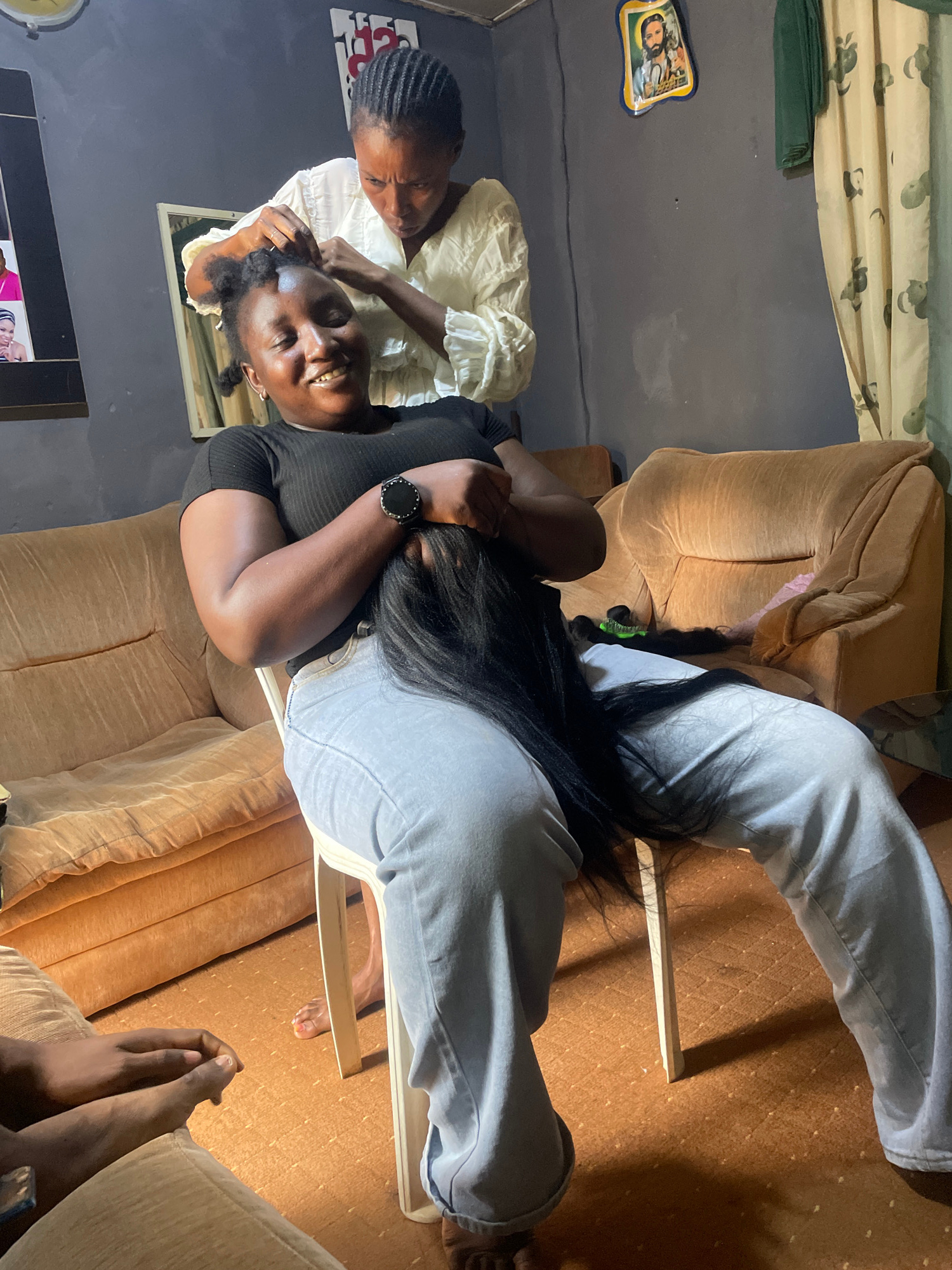 The media file depicts an indoor scene in Jos, Nigeria, likely a home given the casual setting. Two individuals are the primary subjects. One person, wearing a black top and grey pants, is seated and appears to be having their hair braided. They are smiling and looking towards the camera, showing an expression of joy or amusement. The other person, wearing a white blouse, is actively braiding the hair of the seated individual. Their hands are visible, engaged in the braiding process.
The background includes a beige sofa, a dark gray wall adorned with a small framed picture of a man, and a patterned curtain with green and beige tones. The lighting suggests it could be daytime, though artificial lighting might also be in play. The floor is covered with a brown carpet.
Notable details include the long black hair being braided, which is spread out on the seated person's lap and the floor. The seated person is wearing a smartwatch on their left wrist. The overall atmosphere appears relaxed and friendly, with the person being braided showing clear happiness. No text is clearly visible or legible in the provided frames.
