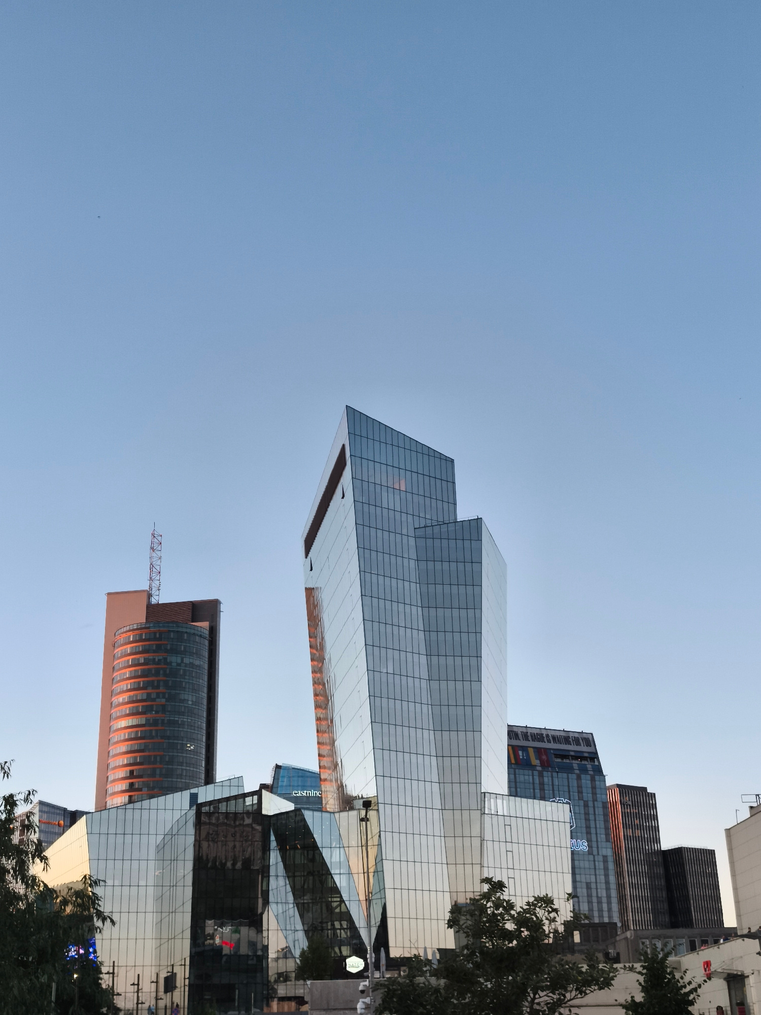 An exterior, low-angle, vertical medium shot captures several modern skyscrapers against a clear, pale blue sky. The buildings are made of glass and metal, with geometric designs. One prominent building is a tall, angular structure with a glass facade reflecting the sky and surrounding structures. To its left is a cylindrical building with orange-tinted glass bands and a metal antenna on top. In the foreground, a lower, angular glass building reflects the sky and other buildings. The scene is set in an urban environment, likely a business district, with hints of greenery at the bottom. No people are clearly visible. The lighting suggests it is either early morning or late afternoon.