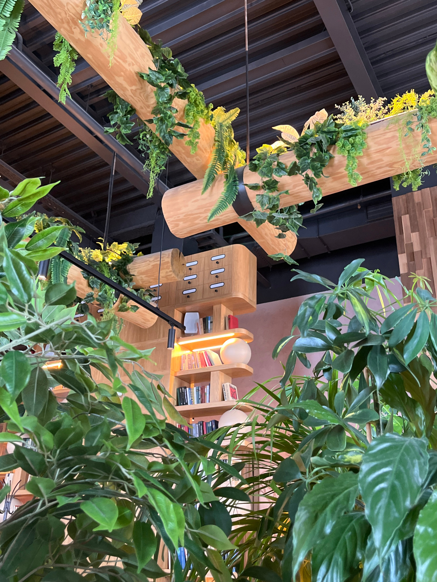 The image captures an interior scene, possibly a cafe or a library, with a strong emphasis on biophilic design elements. Several large, artificial plants with lush green foliage are prominently displayed, some hanging from wooden beams that are suspended from the ceiling. These beams are integrated into the ceiling structure, which appears to be dark and industrial, possibly made of metal.
On the right side of the image, a wooden shelving unit is visible, filled with books. A spherical lamp sits on one of the shelves, emitting a warm glow. Above the shelving unit, another wooden element is mounted, featuring a series of small, white drawers with thin black handles, resembling a miniature filing cabinet or a decorative storage unit.
The background wall is paneled with vertical wooden slats, adding to the natural and organic aesthetic. The lighting in the scene is a mix of ambient light and targeted illumination from the spherical lamp, creating a cozy and inviting atmosphere.
The image is taken from a low angle, looking upwards, which emphasizes the scale of the plants and the suspended wooden structures. No people are visible in the frame. The overall impression is one of a carefully curated and aesthetically pleasing indoor space that incorporates natural elements.
This scene is located in the city of Verkhnyaya Pyshma, Russia, within a commercial establishment.
