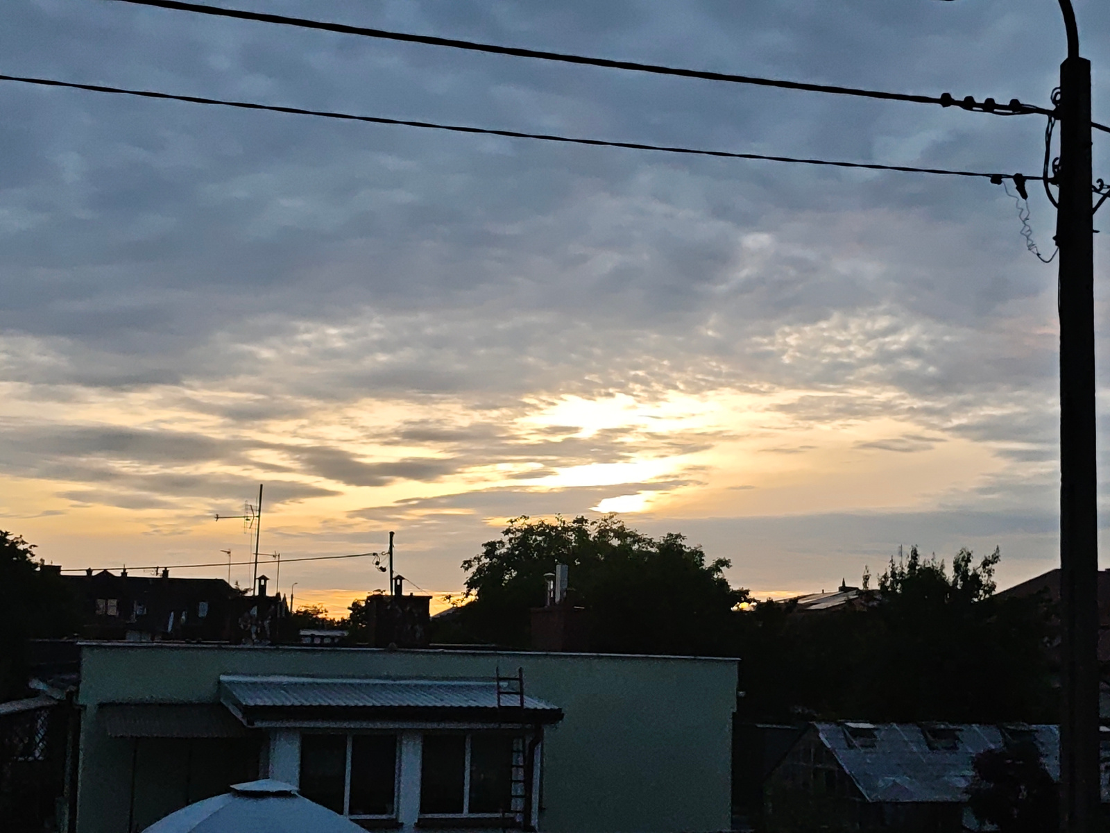 This is a medium-shot, eye-level photograph capturing a view over rooftops in the city of Mława, Poland, during what appears to be sunset. The sky is filled with a mixture of gray and white clouds, with streaks of yellow and orange light breaking through, indicating the sun is low on the horizon. Two dark power lines are prominent in the upper portion of the frame, and a utility pole is visible on the far right, also silhouetted against the sky.
In the foreground and midground, several buildings are visible. The most prominent is a light green building with a corrugated metal roof on the left side. It has a white section at the top and a dark ladder attached to its side. Adjacent to it, other rooftops are visible, some with dark-colored structures and chimneys. Trees are interspersed among the buildings, their dark foliage contrasting with the lighter sky. In the far distance, more rooftops and trees are visible, silhouetted against the colorful sunset. The overall impression is one of a residential or mixed-use urban area at dusk. There are no people or animals visible in the scene.
