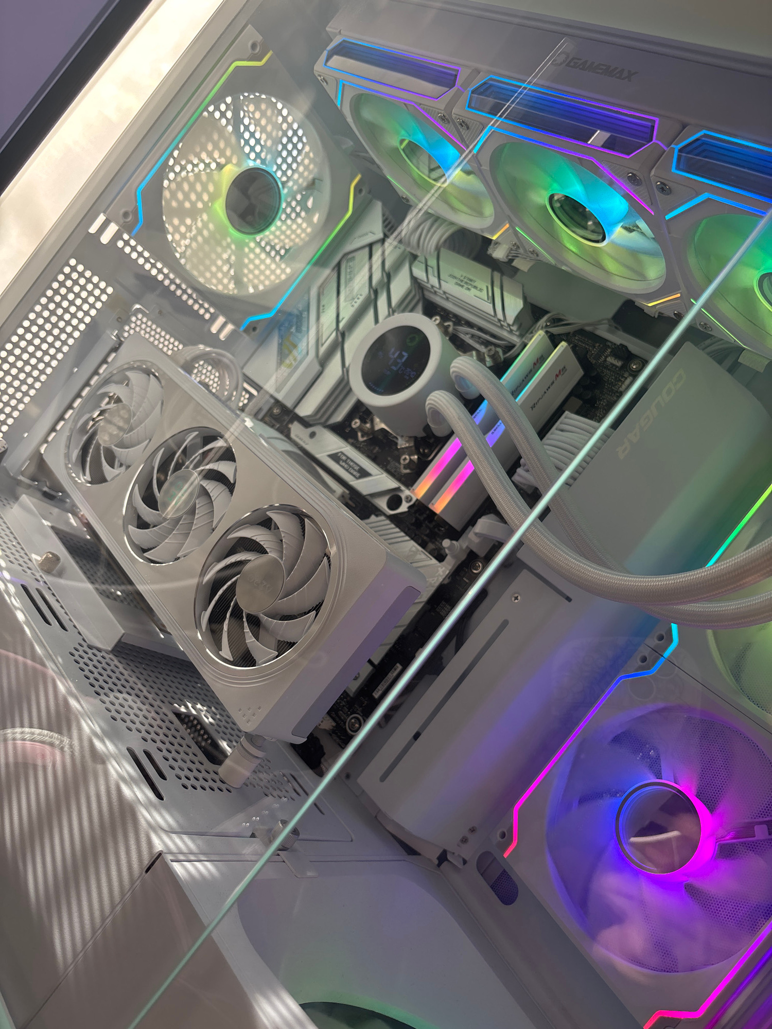 This image shows the interior of a white computer tower with visible components. The case has a glass side panel, allowing a clear view of the internal hardware. Several RGB fans are present, illuminated with various colors including green, blue, and purple, creating a vibrant lighting effect. A large white graphics card with three fans is mounted horizontally, and what appears to be a CPU cooler with a circular display is also visible. The motherboard and RAM modules are also discernible. Cables are neatly managed within the case. The overall impression is of a high-end, aesthetically designed computer system. The location is not discernible from the image.