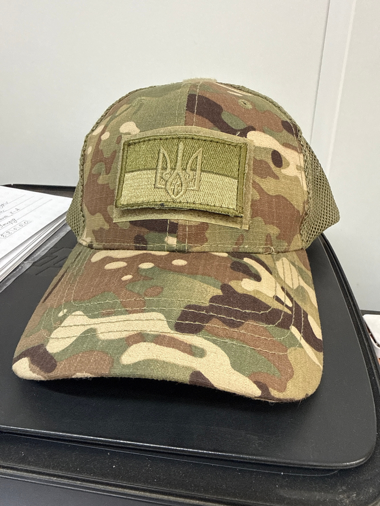 A camouflage baseball cap with a mesh back is displayed on a black surface. A rectangular patch with the Ukrainian trident symbol is affixed to the front of the cap. The background is a plain, light-colored wall.