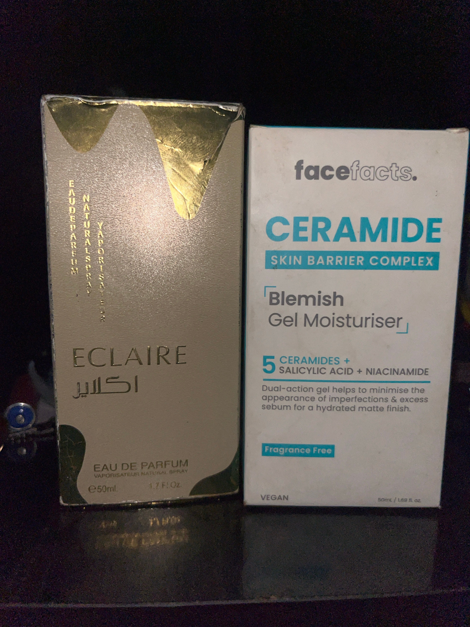 The image displays two product boxes placed on a dark, textured surface, likely a table. The box on the left is a light beige color with metallic gold accents. It features text in gold, including "EAU DE PARFUM" and "ECLAIRE" in large font, with smaller text above and below. The box on the right is white with teal and gray text. It displays the brand "facefacts." prominently at the top, followed by "CERAMIDE SKIN BARRIER COMPLEX" and "Blemish Gel Moisturiser." Below this, there is information about "5 CERAMIDES + SALICYLIC ACID + NIACINAMIDE" and a description of the product's benefits. A "Fragrance Free" label is visible near the bottom. The context of Bukuru, Nigeria is not inferable from the visual information in the image.