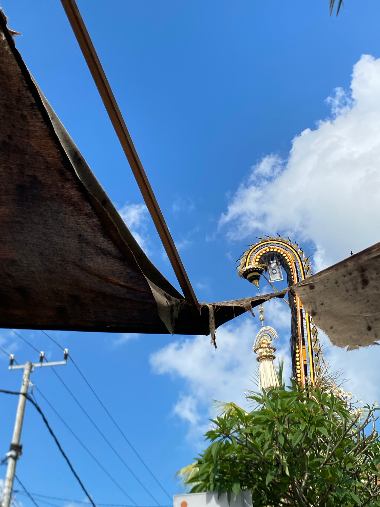This low-angle, eye-level shot captures a vibrant scene under a clear blue sky dotted with white clouds, likely in Mengwi, Indonesia. In the foreground, tattered brown canvas fragments are suspended by wooden beams, casting a shadow and obscuring the view of what's beyond. To the right, a tall, ornate structure, possibly part of a temple or festival decoration, curves upwards, adorned with gold, yellow, and blue accents. A decorative lamp hangs from the structure. Below this, a lush green tree with white flowers is visible. In the distance, a utility pole with wires stands against the sky. The overall impression is one of outdoor, possibly festive, elements against a bright, tropical sky.