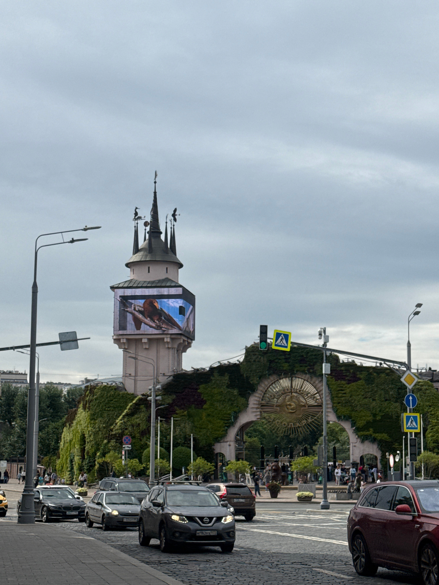The image shows a street scene in Moscow, Russia, featuring the Moscow State Circus (Bolshoi Circus) on Vernadsky Avenue. In the foreground, several cars are driving on a cobblestone street under a cloudy sky. To the left, a tall lamppost with multiple lights stands prominently. In the background, the distinctive tower of the Moscow State Circus is visible, adorned with a large LED screen displaying an image of a bird. The building's facade is covered in lush greenery, forming an archway with a large, ornate golden sun symbol above the entrance. People are seen walking around the entrance and gathered in front of the circus building. Traffic lights and pedestrian crossing signs are also present, indicating a busy urban environment.