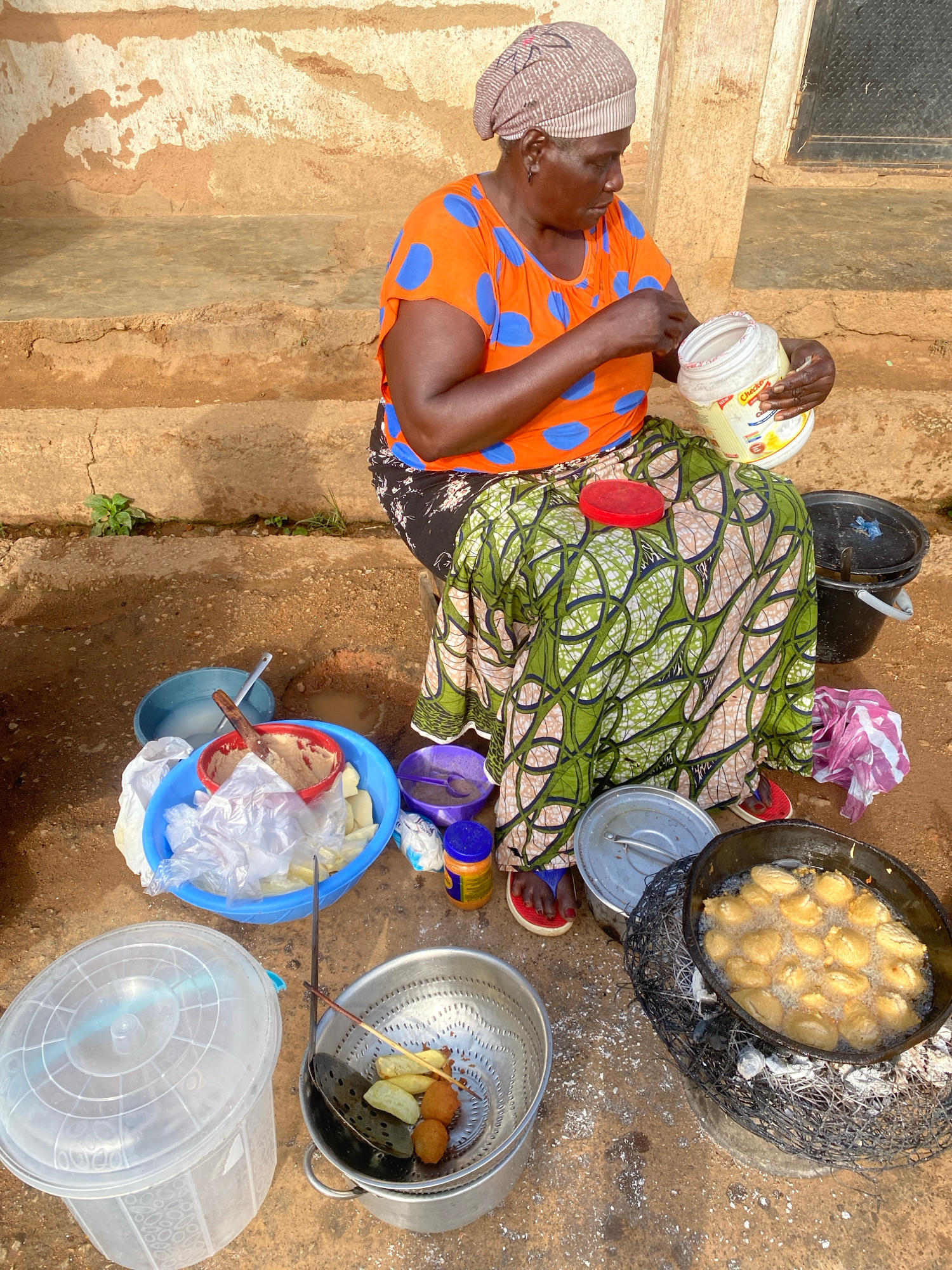 An overhead medium shot captures a woman, likely in Jos, Nigeria, seated outdoors and preparing food. She wears an orange t-shirt with blue polka dots and a patterned wrap skirt. Her head is covered with a beige and brown headscarf. In her right hand, she holds a large white plastic tub with a yellow label, and appears to be adding something from it. In front of her are various cooking implements and ingredients. A large blue bowl contains what appears to be dough and possibly some cut fruit or vegetables, wrapped in plastic. A red bowl with a wooden spoon sits inside the blue bowl. Next to it are smaller purple and blue containers. To the right, a black bucket with a pink and white cloth is visible. A large pan filled with oil and golden-brown fritters is placed over a charcoal brazier, with some of the fritters being fried. A metal colander holds some cooked fritters and skewers. A large clear plastic tub with a lid is situated to the left. The ground is composed of dirt and sand. The background features a rough, textured wall and a dark metal door. The scene suggests a food preparation area for sale or consumption.