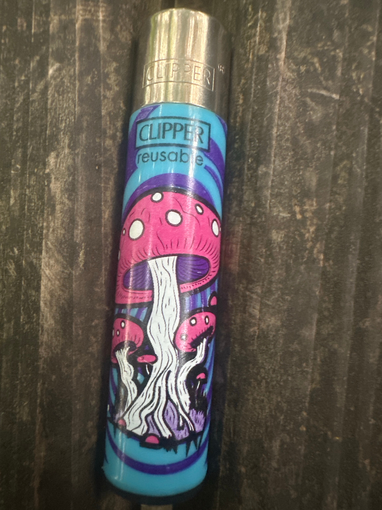 A close-up, overhead, eye-level shot features a Clipper lighter with a psychedelic mushroom design. The lighter is positioned vertically on a dark, textured wooden surface. The lighter's body is predominantly cyan with purple stripes, and it displays a graphic of stylized pink and white mushrooms with purple accents. The word "CLIPPER" is visible in a black box near the top of the lighter, followed by "reusable" in smaller lettering below it. The metal top of the lighter is reflective and shows the "CLIPPER" branding. The background is a dark, vertically grained wood, possibly a table or countertop, with subtle variations in tone and texture. The lighting is even, highlighting the details of the lighter and the surface. The location is identified as Unknown, Unknown.