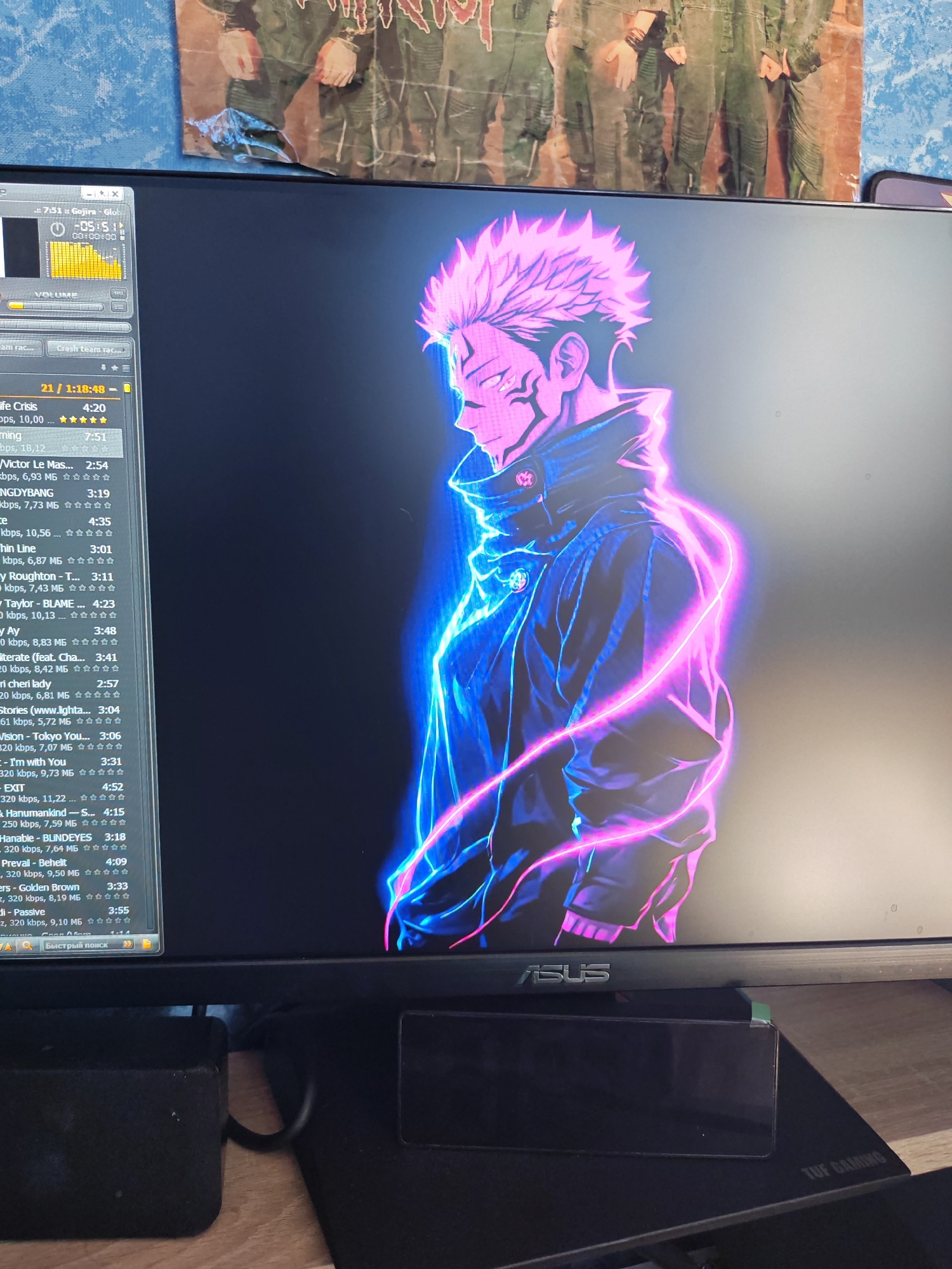 The image displays a computer monitor showcasing a vibrant anime-style wallpaper of a male character with spiky pink hair and glowing blue and pink energy trails around him. The character is wearing a dark jacket. The monitor is positioned on a desk, with a black gaming mousepad and speakers visible below it. In the background, a poster of soldiers in uniform is partially visible.
On the monitor's screen, a media player interface is open, displaying a playlist of songs. Song titles and artist names are listed, along with playback times and bitrate information, such as "Life Crisis 4:20" and "Victor Le Mas... 2:54". The media player has controls for volume and playback. The bottom of the monitor has an "ASUS" logo and a "TUF GAMING" inscription on the mousepad. The overall scene suggests a personal computer setup in a room.