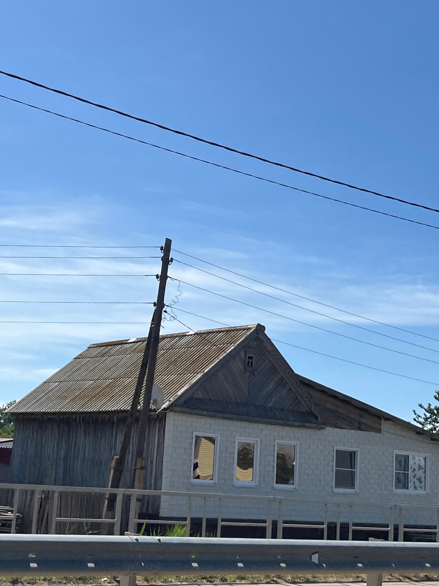 The image depicts a rural scene under a clear blue sky with wispy clouds. In the foreground, a grey metal guardrail runs horizontally across the bottom of the frame. Behind the guardrail, a weathered wooden fence encloses a property. The main subject is a two-story house with a corrugated metal roof. The left side of the house appears to be clad in vertical wooden planks, while the right side is constructed of light-colored brickwork. Several windows are visible on the brick section of the house, some with curtains. A wooden utility pole with multiple wires attached stands prominently to the left of the house, with a satellite dish mounted on its upper section. The overall impression is that of a somewhat aged but functional dwelling in a non-urban setting.