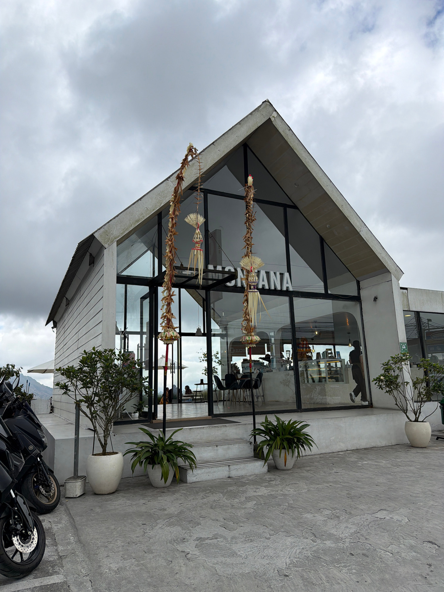 The image depicts a modern, angular building with large glass windows, likely a cafe or restaurant, situated in Kintamani, Indonesia. The building's exterior is a mix of white siding and concrete. Two tall, decorative Balinese-style hanging ornaments, made of dried palm leaves and straw, are suspended from the eaves of the building, just outside the entrance.
In the foreground, to the left, are three black motorcycles parked on a concrete surface. The area in front of the building is paved with concrete. Several potted plants, including a small tree and a leafy bush, are strategically placed around the entrance and along the building's facade.
Inside the building, visible through the glass, are tables and chairs, and a counter area, suggesting a commercial establishment. Several people are seated inside, and one person is walking near the entrance. The sky is overcast with clouds, and a mountainous landscape is faintly visible in the background, indicative of Kintamani's elevated location. The overall atmosphere is somewhat subdued due to the cloudy weather.