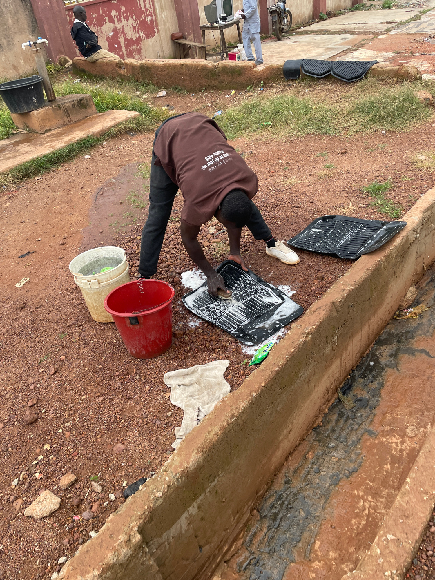 The image depicts an outdoor scene on a dusty, reddish-brown ground, possibly in the city of Jos, Nigeria. In the foreground, a young man wearing a brown t-shirt with text and dark pants is actively scrubbing two black car floor mats with a brush and soap. The mats are covered in white suds. To his left are two buckets, one a lighter color and the other red, with one appearing to contain water and green items. A white cloth lies on the ground near the buckets. In the background, a young man in a black jacket and light-colored pants is seated on a low concrete wall, facing away from the viewer. A water tap is visible on a concrete pillar near a large black bucket. Further back, a table with electronic equipment and a motorcycle can be seen. The environment suggests an informal setting, possibly a residential area or a car wash service. A concrete drainage channel runs along the right side of the image, filled with murky water and debris.