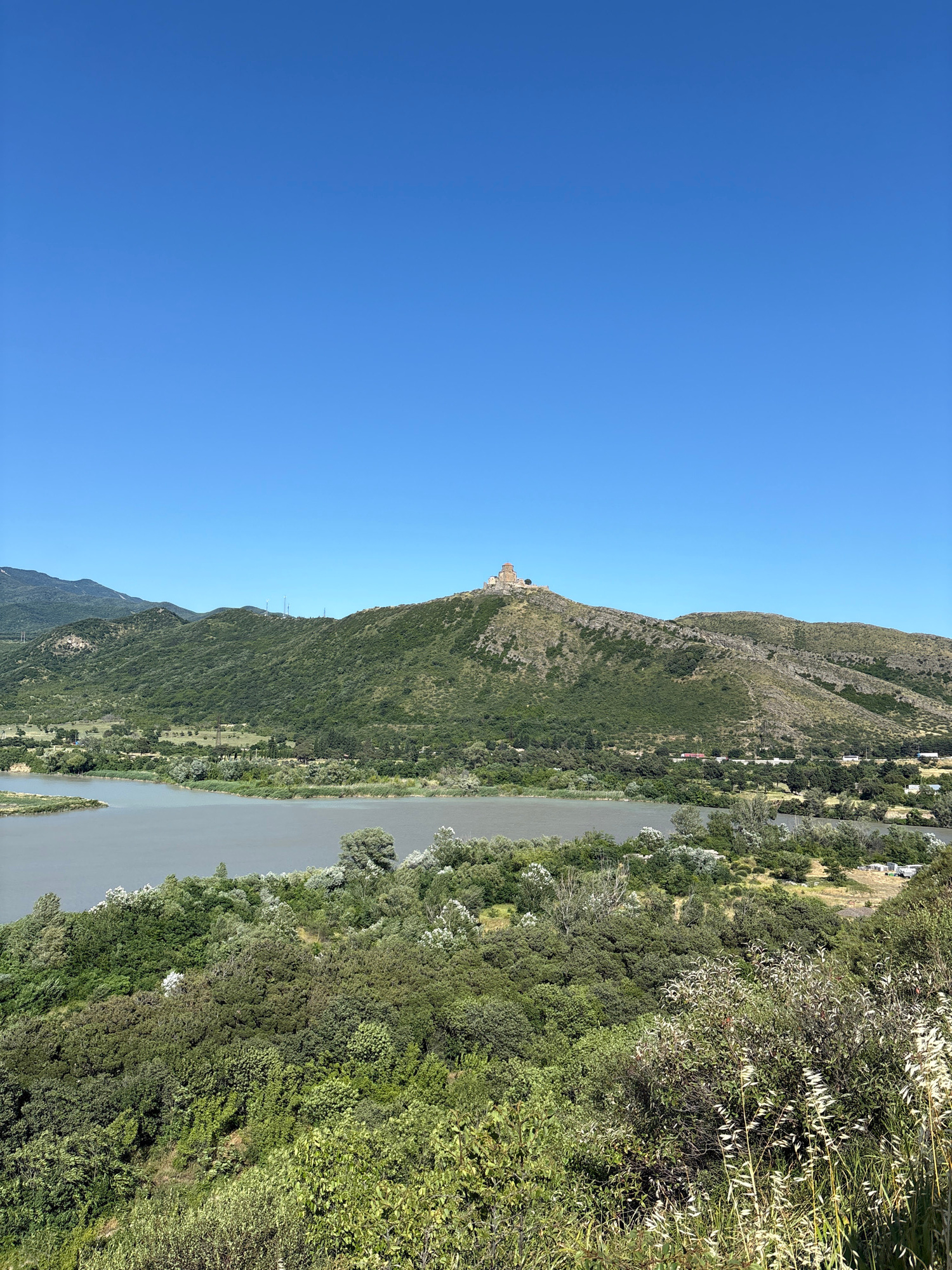 A wide shot captures a serene landscape on a clear, sunny day. A vast body of water, likely a river or lake, dominates the left foreground, its surface a muted gray. Lush green foliage, a dense mix of trees and shrubs, lines the banks and extends up the slopes of rolling hills that fill the middle ground and background. The hills are covered in verdant vegetation, with rocky outcrops visible on some of the steeper inclines.
Perched atop the highest hill, in the center of the frame, is a historic stone structure, a monastery or church, its weathered stone contrasting with the vibrant green surroundings. The structure has a distinctive dome and appears to be ancient. The sky above is an unbroken expanse of brilliant blue. The overall impression is one of natural beauty and historical significance.
This scene depicts the Jvari Monastery in Mtskheta, Georgia, situated on a hilltop overlooking the confluence of the Aragvi and Mtkvari rivers.
