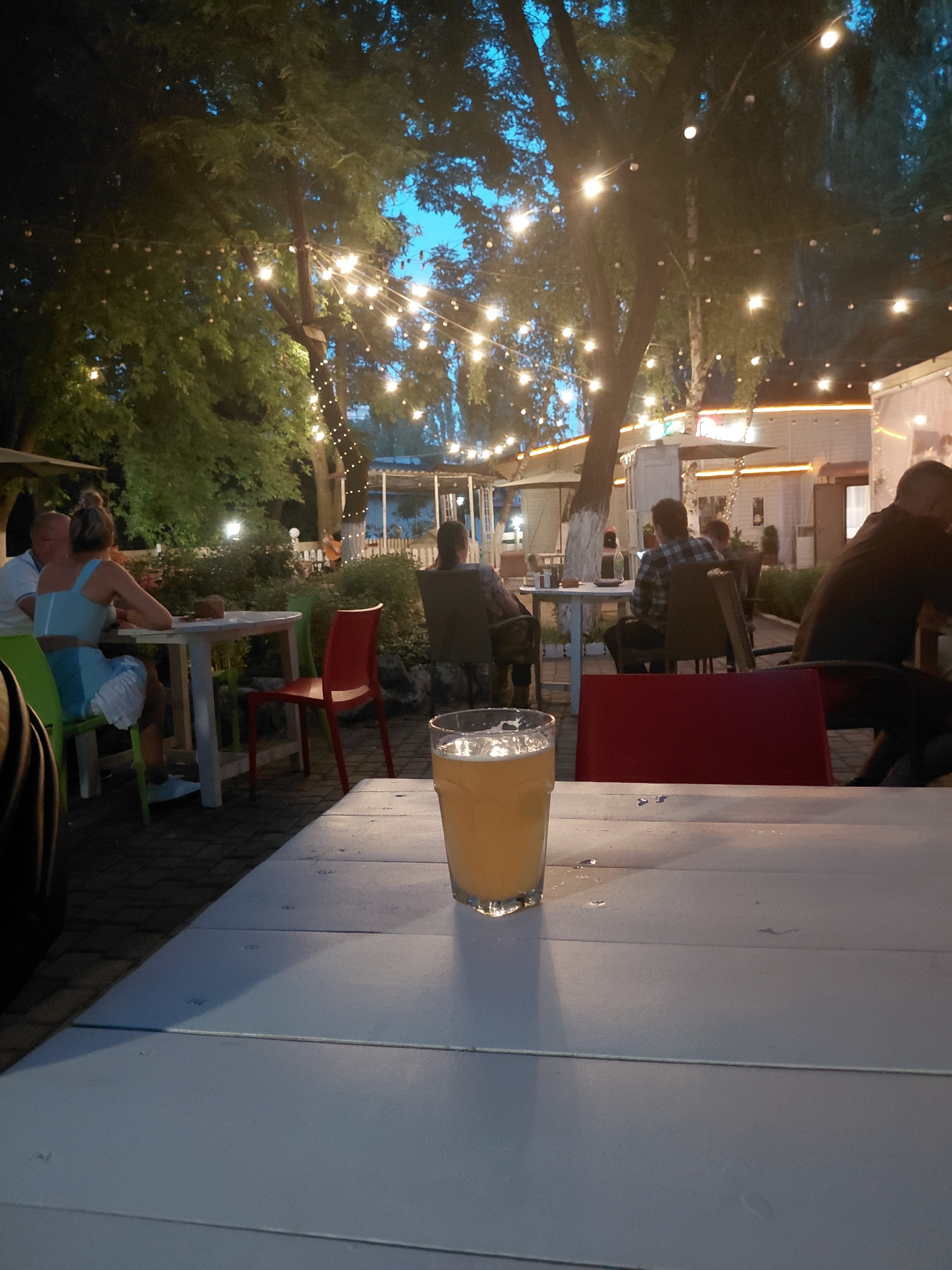 The image depicts an outdoor dining scene at an establishment with string lights overhead, set in the evening or twilight hours. In the foreground, a white table occupies the lower portion of the frame, with a glass of a yellow beverage placed on it. Further back, patrons are seated at various tables. To the left, a man and a woman are seated at a white table, with the woman wearing a light blue dress. To the right, several individuals are visible, including a man in a checkered shirt sitting at a table with a laptop, and another person in dark clothing. The environment appears to be a patio or garden area, with trees and foliage contributing to the ambiance. The lighting from the string lights and possibly from the establishment itself creates a warm, inviting atmosphere.
