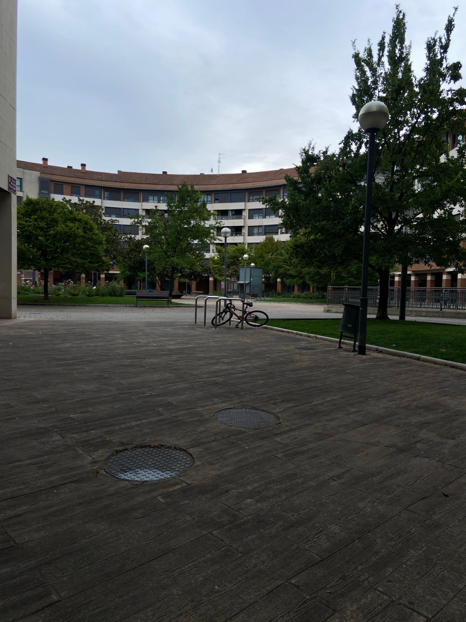 The image depicts an outdoor urban scene in Logroño, Spain, featuring a curved building with multiple stories, lined with windows and topped with a terracotta-tiled roof. Trees with dense green foliage surround the building, interspersed with streetlights. In the foreground, a paved area made of dark wooden planks exhibits two manhole covers. A bicycle is parked near a metal rack, and a dark trash can stands next to a lamppost. The sky is overcast and pale blue. The scene appears to be a public walkway or plaza, possibly within a residential complex or park area, as suggested by the presence of greenery and the design of the building.