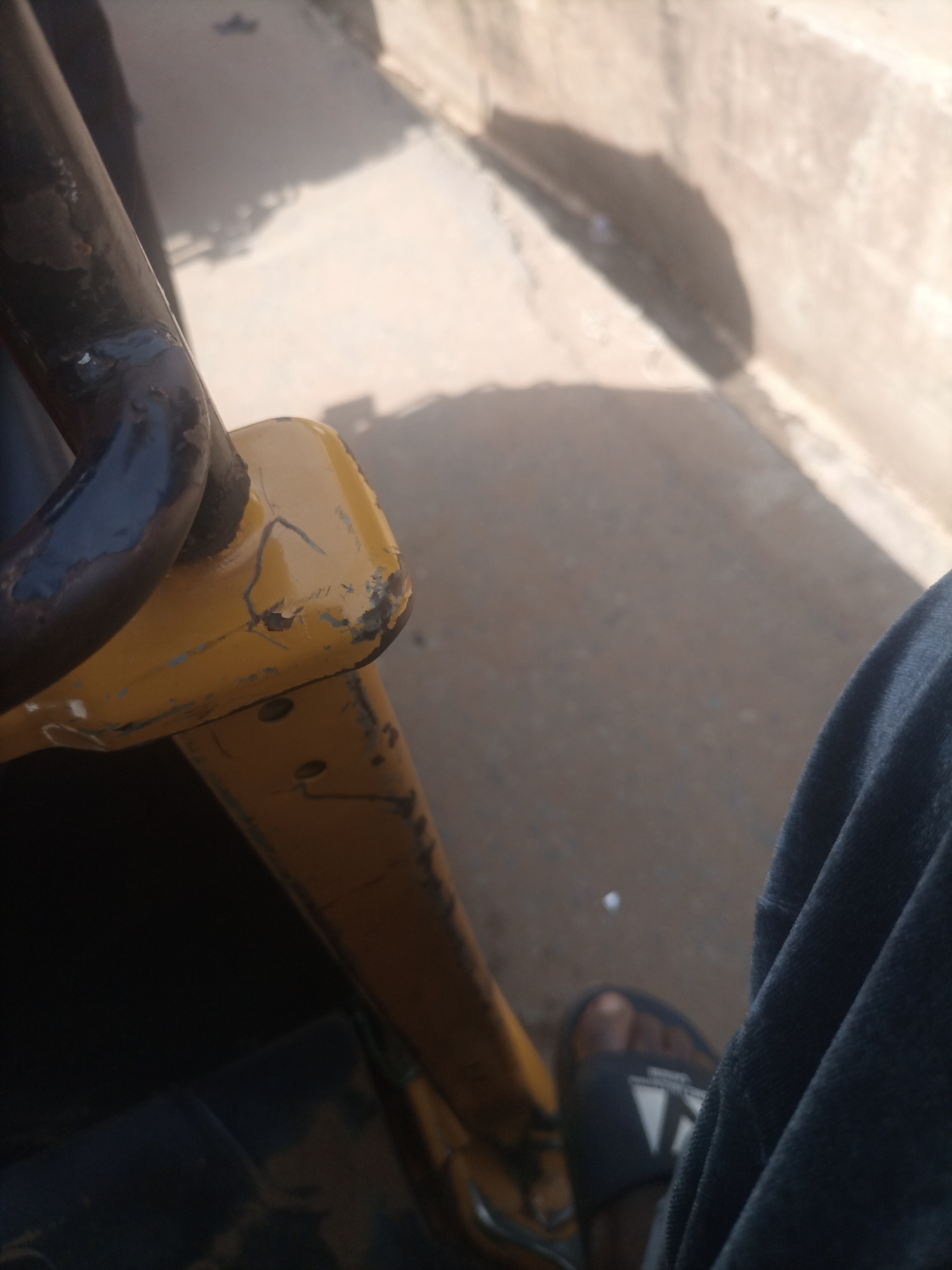 The image captures a close-up perspective from within a vehicle, likely a motorized tricycle or tuk-tuk, in Nkpor, Nigeria. The focus is on a section of the vehicle's yellow-painted frame, which shows signs of wear and tear, including chipped paint and visible metal underneath. A dark, curved metal bar, possibly part of the vehicle's structure or a handrail, is also prominent. In the lower right of the frame, a person's foot wearing a dark sandal with white markings is visible, resting on the vehicle's floor. The person's dark blue or black clothing is also visible. The background consists of a blurred, outdoor environment, suggesting a street or paved area, with shadows cast by an unseen object, likely the vehicle or its surroundings. The overall impression is an ordinary, everyday scene of transportation in a Nigerian city.