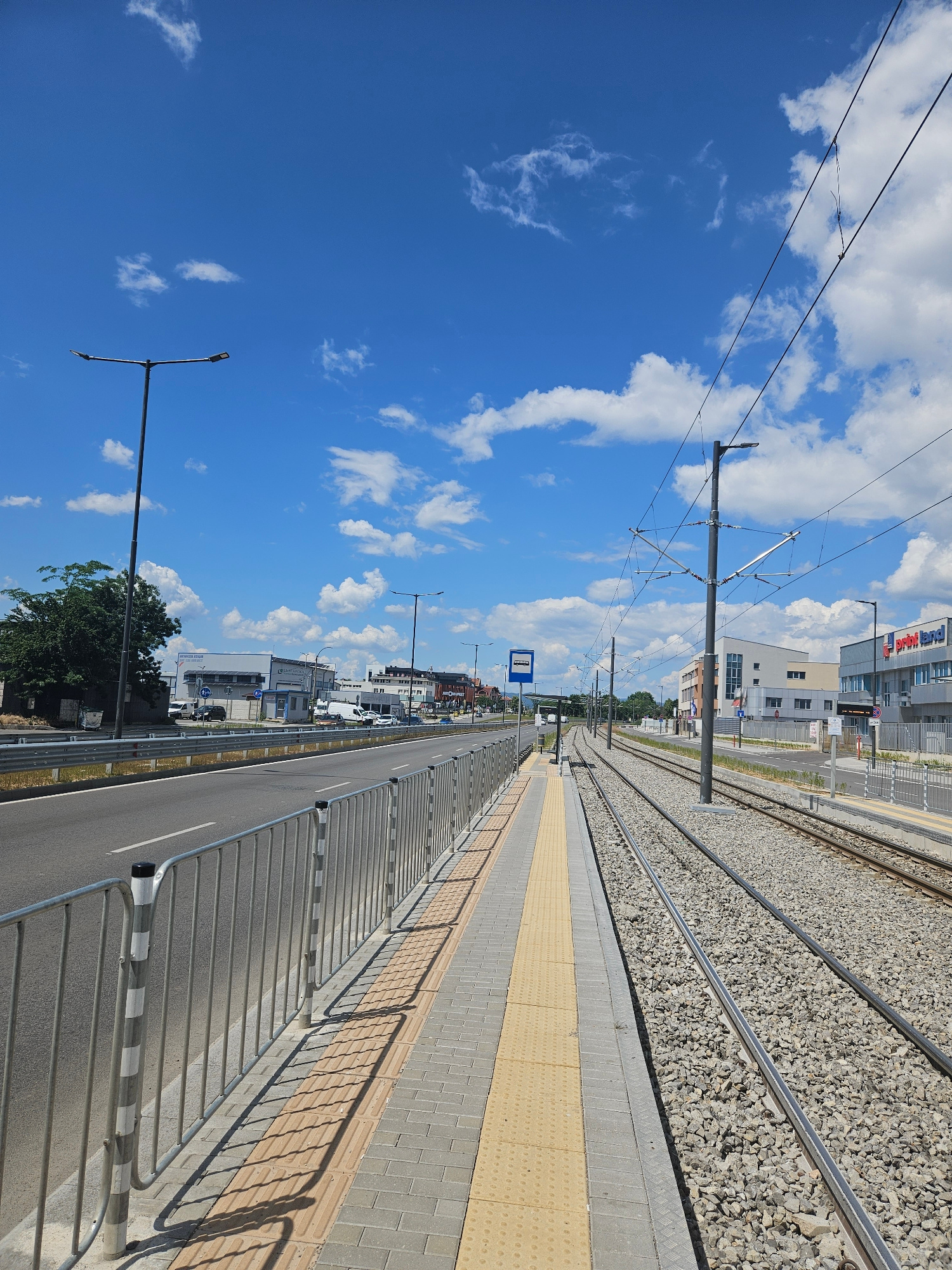 This image depicts a modern tram or train station platform in Sofia, Bulgaria, on a bright, sunny day with a clear blue sky dotted with fluffy white clouds. The perspective is looking down the length of the platform, which is paved with grey bricks and features a yellow tactile paving strip for visually impaired passengers. A metal fence with a patterned railing runs along the edge of the platform, separating it from the adjacent road.
The tram tracks, laid on a bed of grey gravel, run parallel to the platform. Overhead, electrical wires for the tram system are visible, supported by tall, grey poles. In the background, several modern buildings are visible, including what appears to be a commercial establishment with signage. A blue sign indicating a bus stop is also present.
The scene is quiet, with no people visible on the platform, suggesting it might be outside of peak hours. The lighting indicates it is daytime, likely late morning or early afternoon, given the strong shadows cast by the fence and railing. The overall impression is one of an organized and functional urban transit point. No specific text other than the bus stop sign is clearly legible in the image.