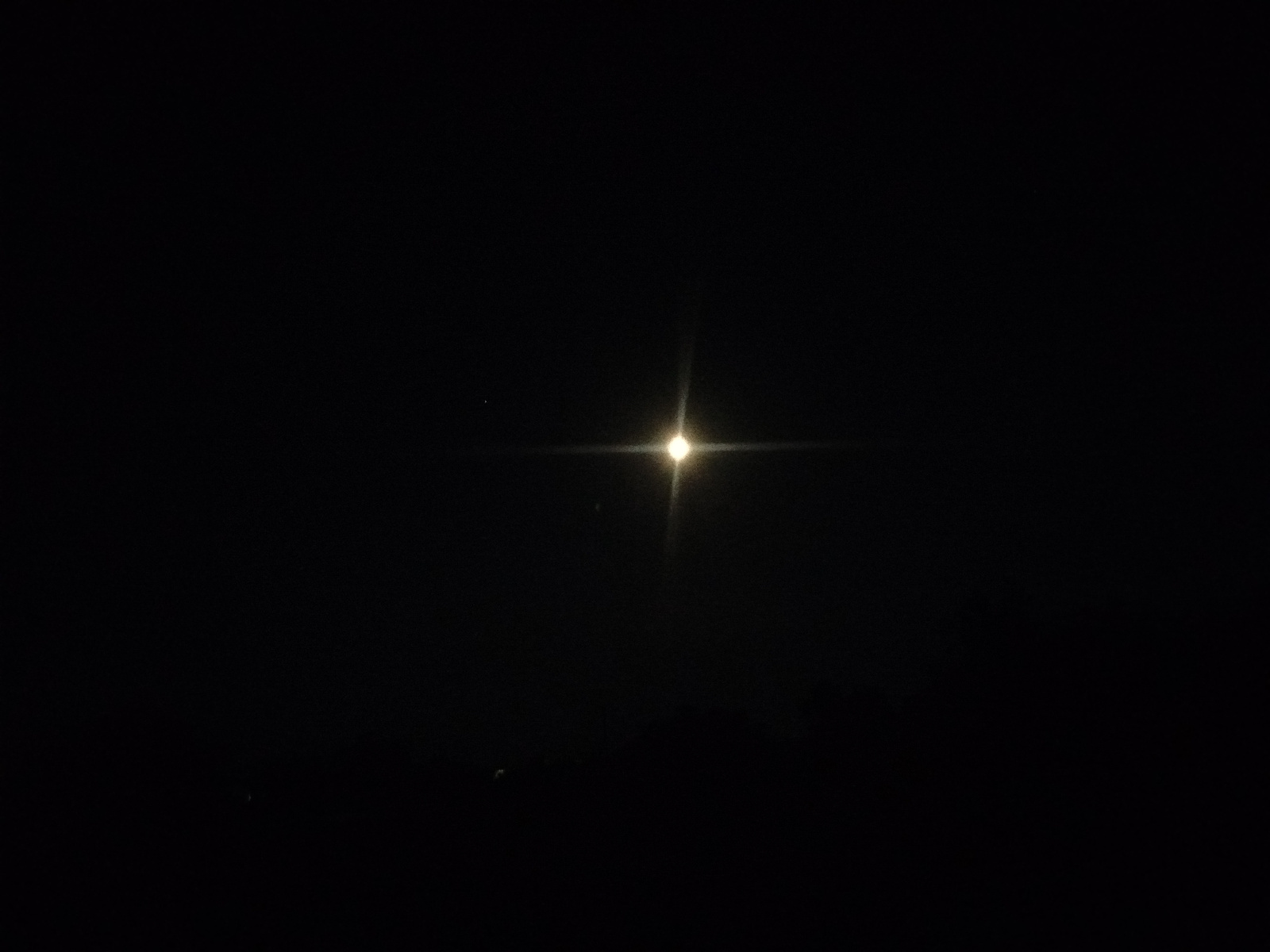 A nighttime, long-exposure photograph captures a bright, circular light source in the center of the frame, creating a starburst effect with four distinct points of light extending horizontally and vertically. The background is a deep, inky black, suggesting a night sky with minimal ambient light. To the left of the central light source, a much smaller, fainter point of light is visible. The overall impression is of a celestial body or a distant artificial light against a dark, possibly cloudless sky. No people, objects, or specific location cues are discernible, beyond the implication of an outdoor setting at night. The image is likely taken in Kostiantynivka, Ukraine, based on the provided context. There is no visible text in the image.