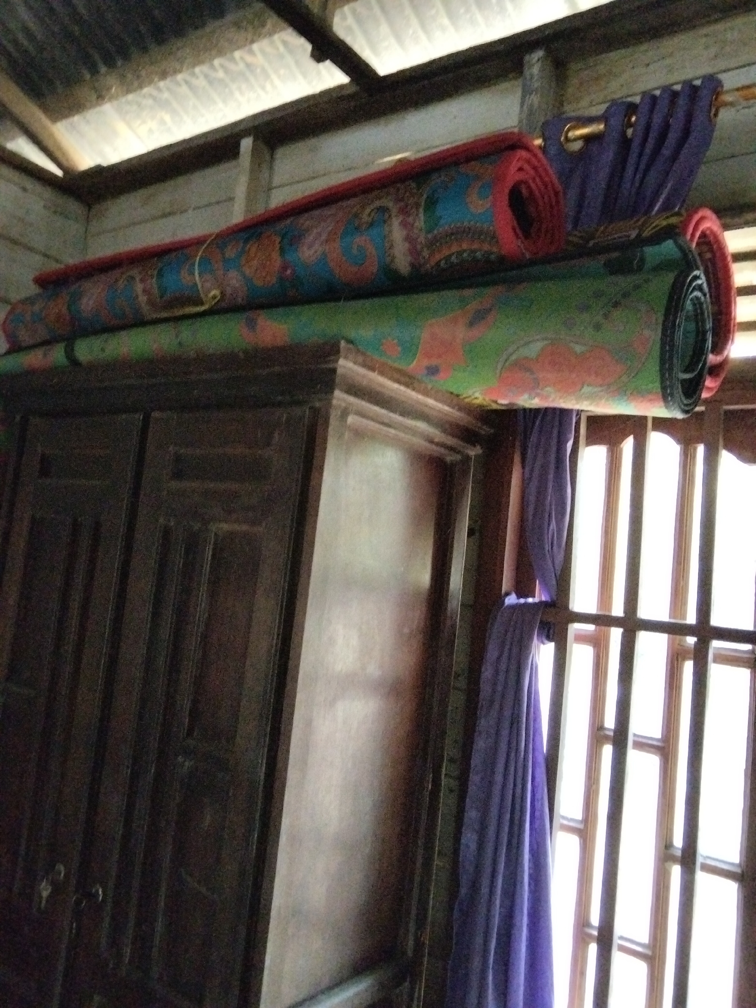 The image shows a rustic interior scene, likely a room or storage area, featuring a dark wooden cabinet. Rolled-up carpets with colorful paisley-like patterns are stacked on top of the cabinet, with the top carpet having a red edge and the bottom carpet featuring green and orange designs. To the right of the carpets, a purple curtain hangs from a rod, obscuring a wooden-framed window. The ceiling appears to be made of corrugated metal, supported by wooden beams. This scene is not explicitly identifiable as being in Benuaraja, Indonesia, as there are no discernible landmarks or identifying features specific to that city.