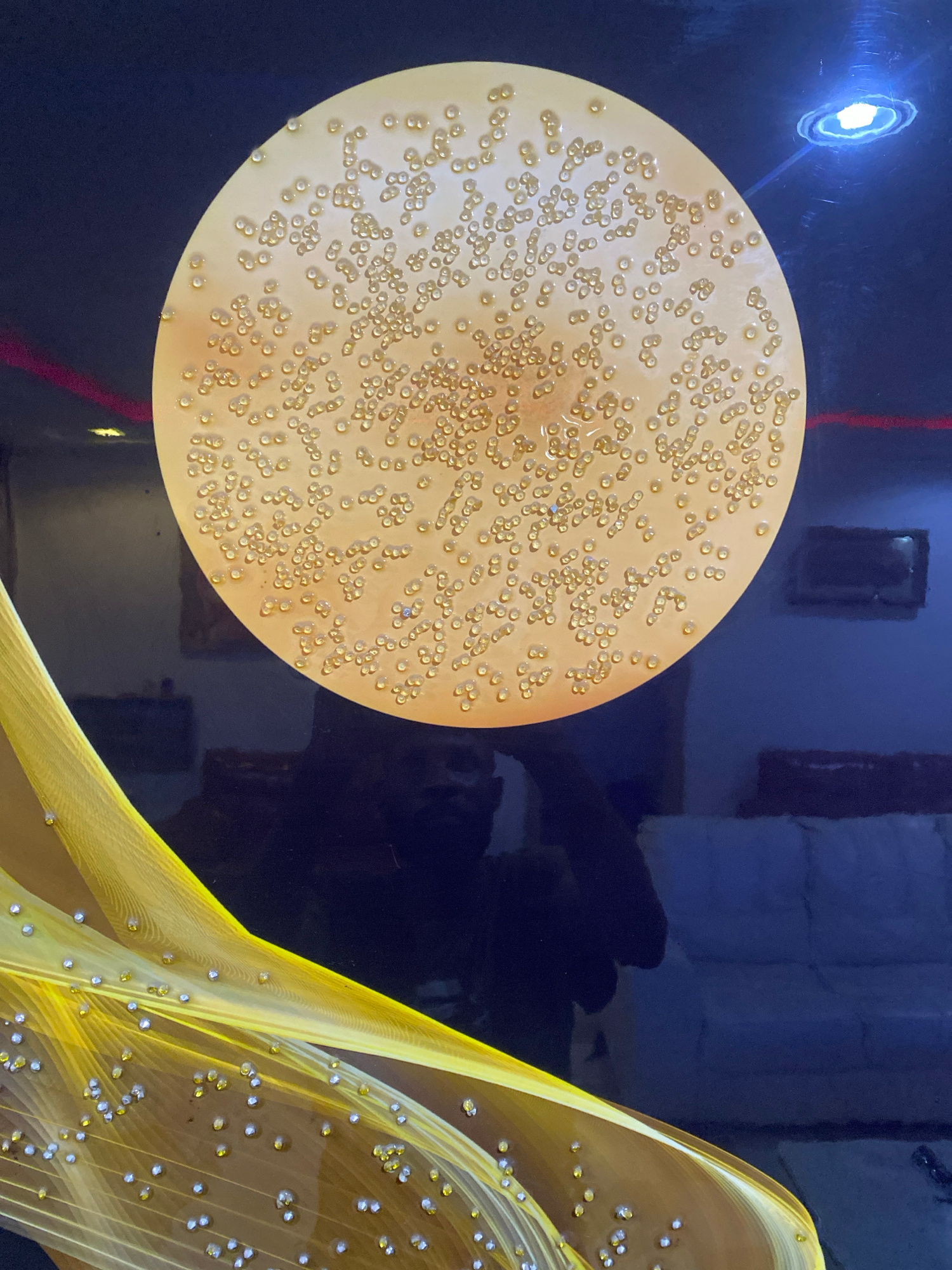A close-up, low-angle shot captures a framed artwork featuring a large, circular, light-yellow disc adorned with numerous small, raised, translucent yellow dots, resembling a magnified biological specimen or a patterned textile. The disc is set against a dark blue background, which reflects ambient light sources, including a bright white light in the upper right corner with a blue halo, and a faint red light along the left edge. In the lower left quadrant, a flowing, golden, translucent fabric-like element, possibly part of the artwork, is heavily embellished with scattered clear and yellow beads. The dark background also shows reflections of what appear to be furniture, possibly couches, and the indistinct silhouette of a person, likely the photographer. The overall scene suggests an indoor setting, potentially a gallery or a decorated room. There is no visual information within the image to suggest the location is Giginyu, Nigeria.