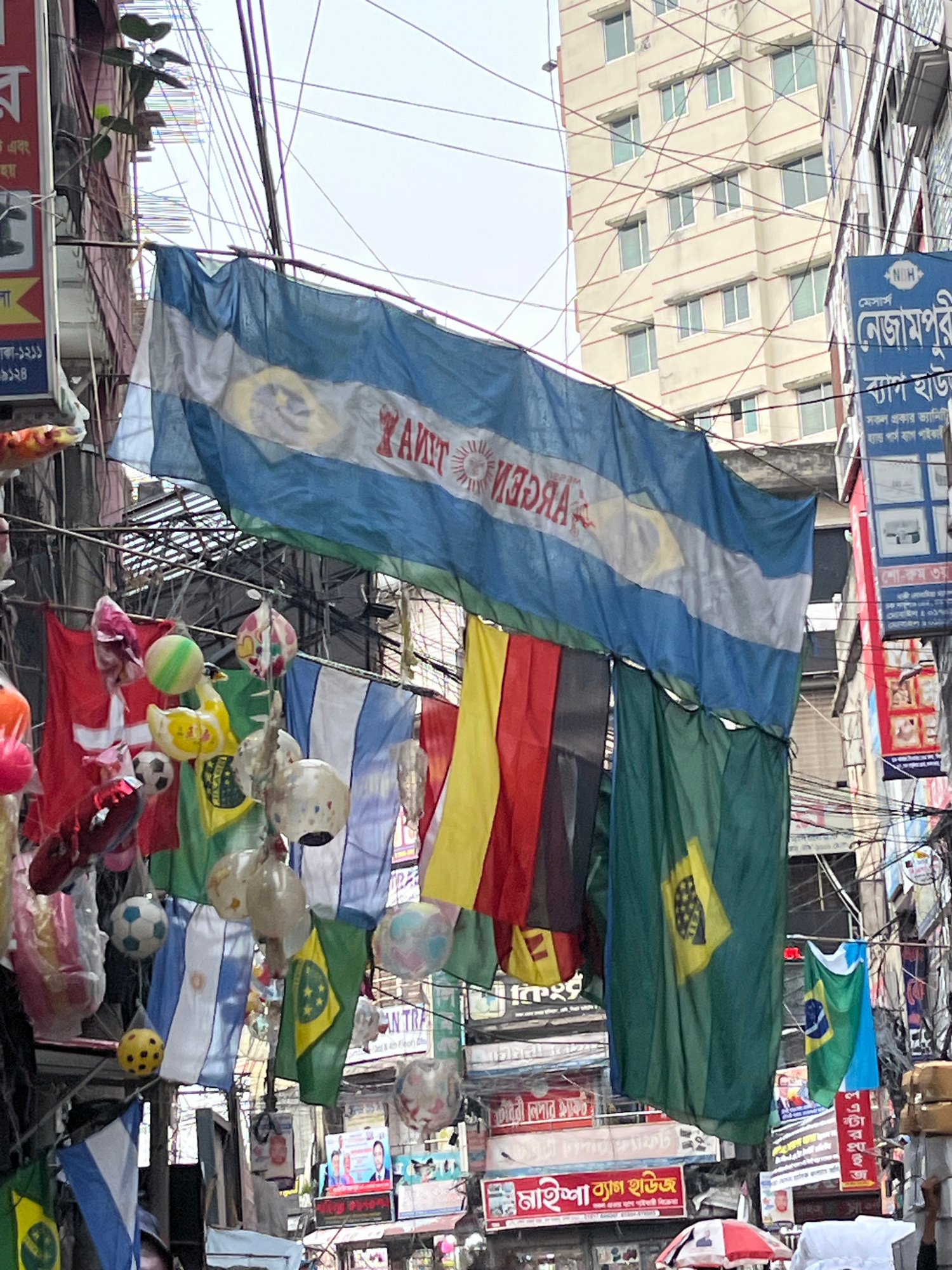The image captures a vibrant street scene in Dhaka, Bangladesh, likely in a commercial area based on the dense signage and overhead wires. The focal point is a collection of national flags, primarily Argentina and Brazil, strung across the street, suggesting a sporting event or national celebration. Among the flags, various decorative items are suspended, including footballs, inflatable toys like a yellow duck, and spherical ornaments, adding a festive and somewhat chaotic atmosphere. Buildings with multiple floors and numerous windows form the backdrop, their facades obscured by a dense network of electrical and telecommunication wires. Signs in Bengali adorn the buildings, indicating local businesses. In the lower portion of the image, people are visible, some partially obscured, suggesting a bustling public space. The overall impression is one of a lively, densely populated urban environment.