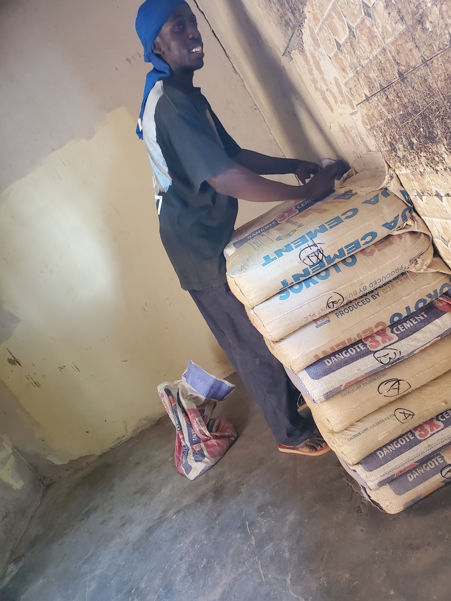 This media file captures a person in a construction or storage setting, likely in Bukuru, Nigeria. The main subject is a person wearing a dark t-shirt, blue head covering, and dark pants, with a white garment peeking out from underneath their shirt. They are positioned in the lower left of the frame, appearing to be interacting with a stack of cement bags. The person's face is visible, and they have a slight smile.
The scene is dominated by several large bags of cement, neatly stacked against a wall. The bags are light-colored with prominent blue and red lettering. Visible text on the bags includes "DANGOTE CEMENT," "BY BUA," and "NT PLC." Some bags also have markings like "A" and "B" drawn on them, possibly for inventory or organization. One bag mentions "CHLORIDE LIMIT: CHLORIDE < 0.10%." Another bag has "PRODUCED BY BUA CEMENT" printed on it.
The background consists of a tiled wall with some discoloration and what appears to be rough concrete flooring. A crumpled, striped bag (red, white, and blue) lies on the floor to the right of the cement bags. The lighting suggests it's daytime, and the overall environment seems to be an indoor storage area or a partially finished construction site. The image is oriented vertically, with the person and cement bags running from top to bottom. The activity depicted is the handling or organizing of cement bags, with the individual actively engaged with the materials.