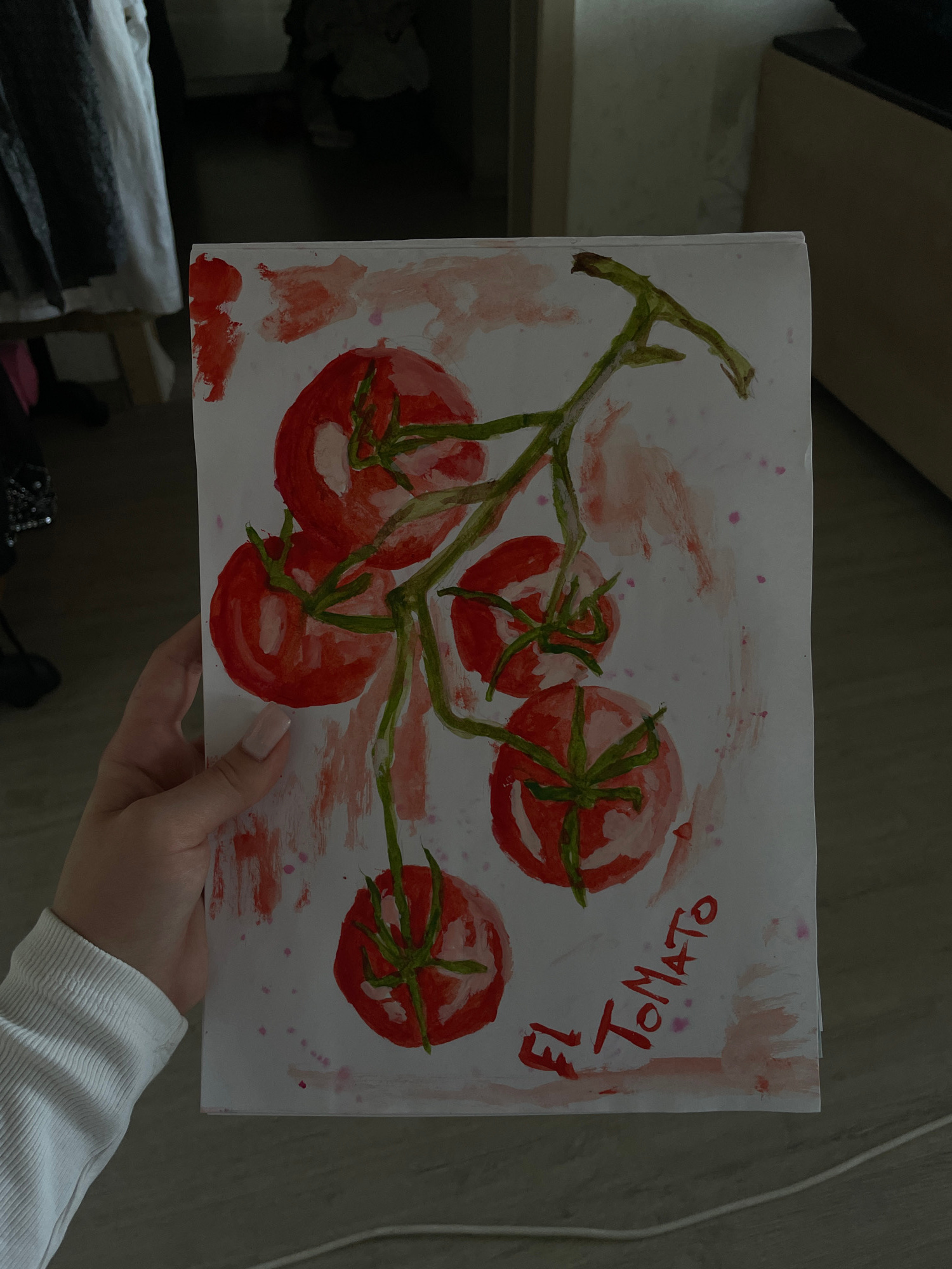 A person's hand, with light skin and short, light-colored fingernails, is holding up a watercolor painting of a vine with several red tomatoes. The painting is on white paper and features green stems and leaves. There are red and pink splotches and splatters on the paper, and the words "EL TOMATO" are written in red paint on the bottom right. The background is dark and out of focus, suggesting an indoor setting with wooden flooring and some clothing on a rack to the left. The image does not contain any recognizable landmarks or elements that would allow for its location to be determined in Minsk, Belarus.