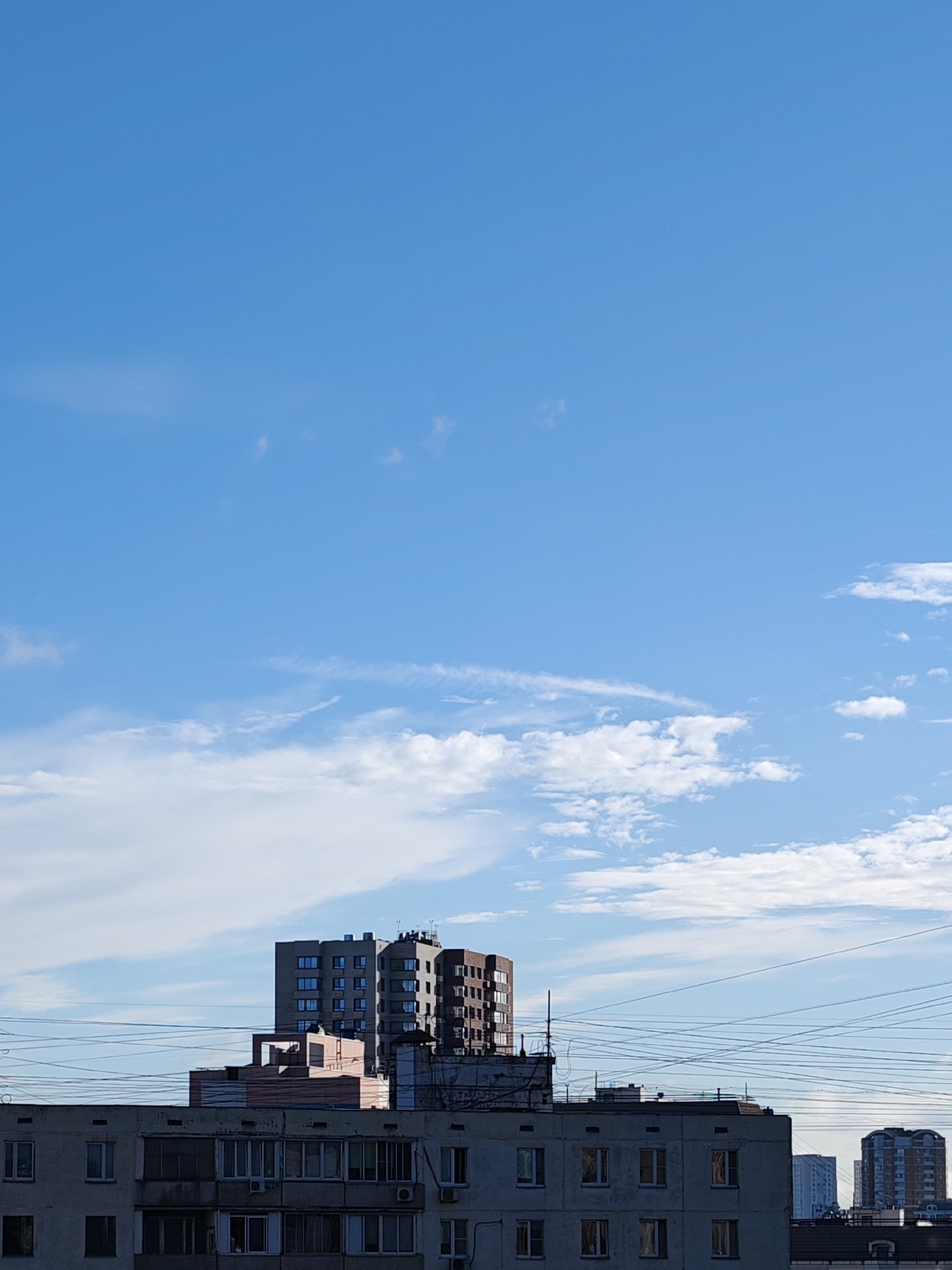 An eye-level outdoor medium shot shows a clear blue sky with sparse white clouds above a cityscape. The cityscape consists of several multi-story buildings of various architectural styles and colors, with numerous power lines crisscrossing the scene. In the foreground, a grey, weathered apartment building with visible windows and air conditioning units stretches horizontally across the frame. Behind it, a cluster of three modern, tall apartment buildings with multiple windows are visible. Further in the background, other buildings of different heights and shapes can be seen. The overall lighting suggests it is daytime.