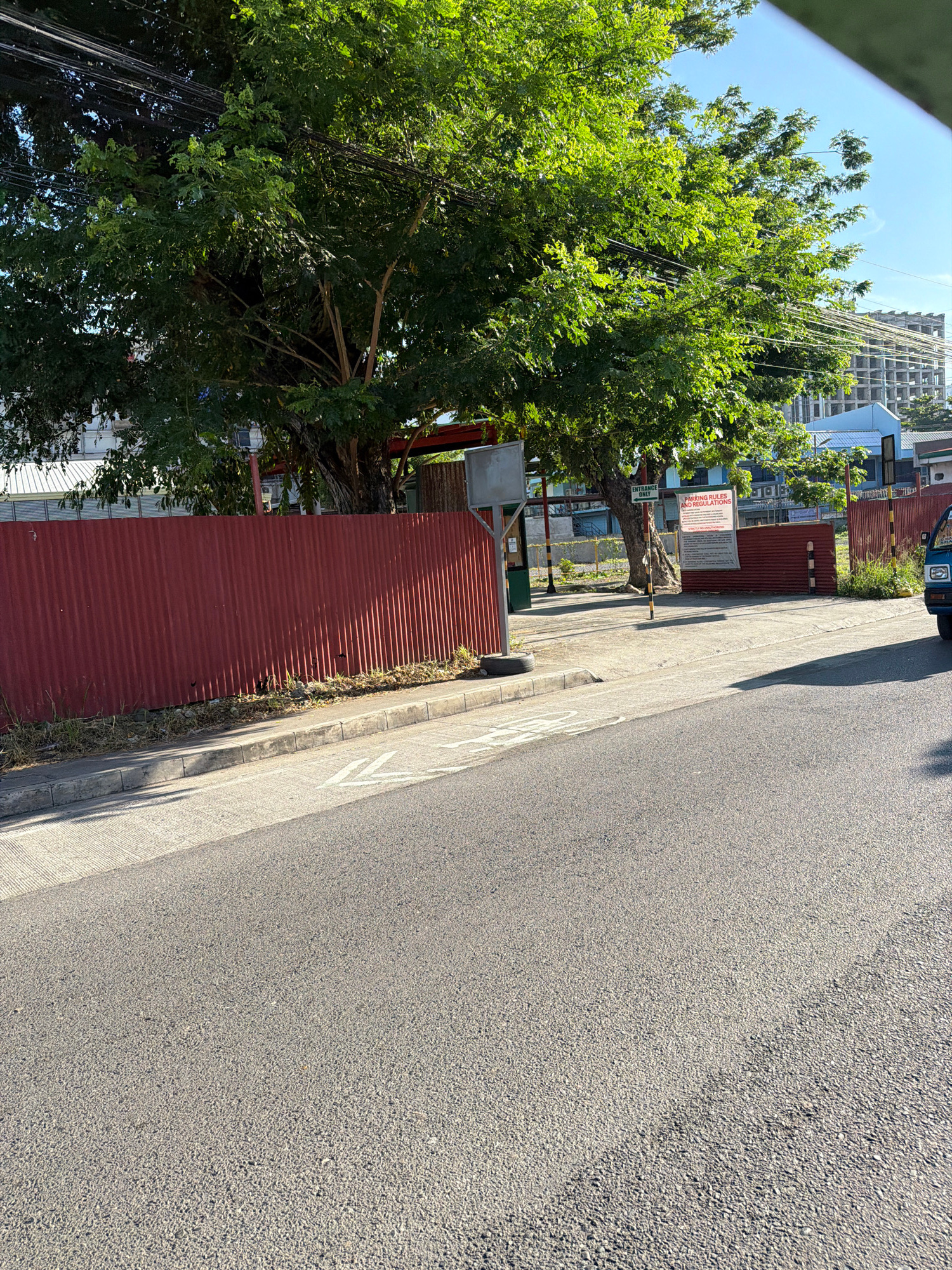 In Davao City, Philippines, a wide asphalt road runs parallel to a corrugated red metal fence. Behind the fence, a large, leafy tree with visible power lines overhead casts shadows on the paved ground. Beyond the tree, a building under construction, with visible concrete pillars and floors, dominates the background. A blue and white building is also visible. Signs are posted near the entrance to the property, with one prominently displaying "PARKING RULES AND REGULATIONS." A blue vehicle is partially visible on the right side of the image. The sky is clear and blue.