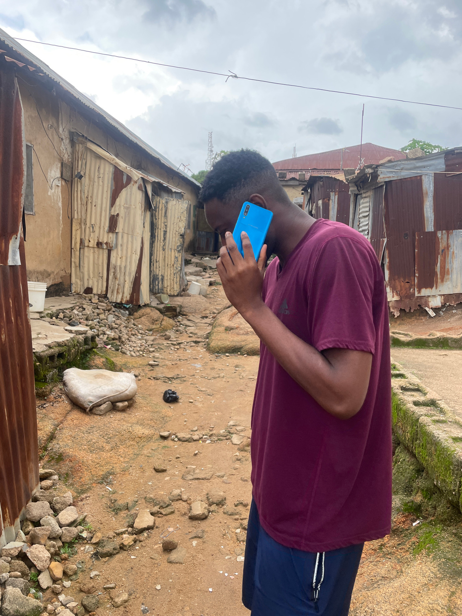 A person is holding a bright blue smartphone to their ear, as if talking on the phone. They are wearing a maroon t-shirt and dark blue shorts. The person is standing in an outdoor environment that appears to be a low-income area. In the background, there are structures made of corrugated metal and concrete, suggesting a somewhat informal settlement. The ground is dusty and uneven, with scattered rocks and some sparse vegetation. The sky is overcast with clouds, indicating a cloudy day. The overall scene depicts a common, everyday moment of communication amidst a rustic, possibly urban, setting. There is no visible text in the image. The time of day appears to be daytime due to the lighting, though the overcast sky makes it difficult to determine a specific time. The location cues, such as the building materials and the overall environment, are consistent with a place like Jos, Nigeria. There are no strong emotional cues visible from the person, as their face is partially obscured.