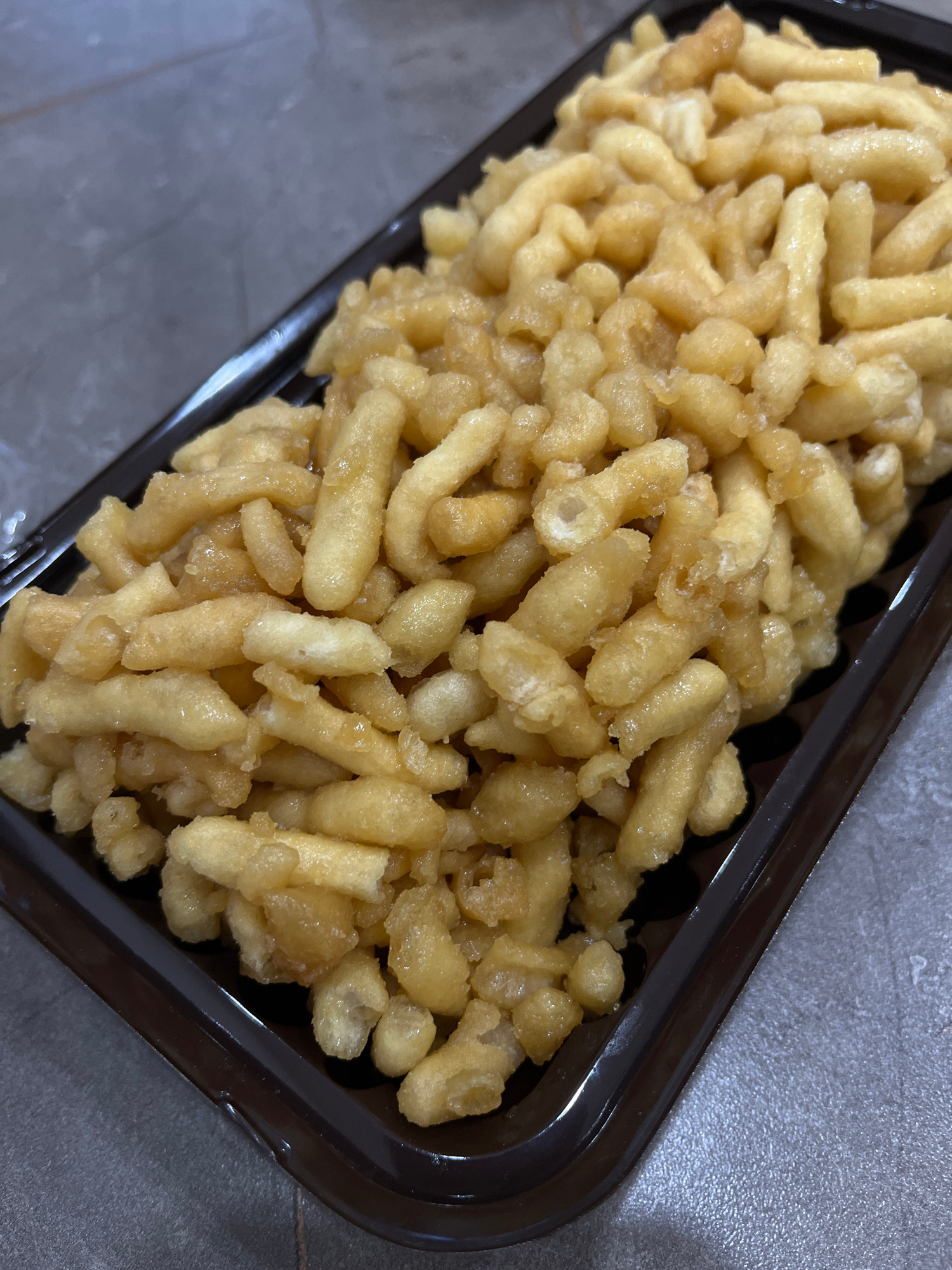 The image shows a close-up, overhead view of a rectangular, dark brown plastic container filled with a golden-brown, irregularly shaped fried pastry, likely a type of sweet or dessert. The pastries appear to be coated in a syrup, giving them a glossy sheen. They are densely packed within the container. The container itself is placed on a textured, gray surface that resembles a countertop or table. The background is out of focus and dark. There are no people or animals visible in the image. The context suggests this might be a food item prepared or sold in Novokuybyshevsk, Russia, though no specific landmarks or identifying features of the city are present in the image itself.