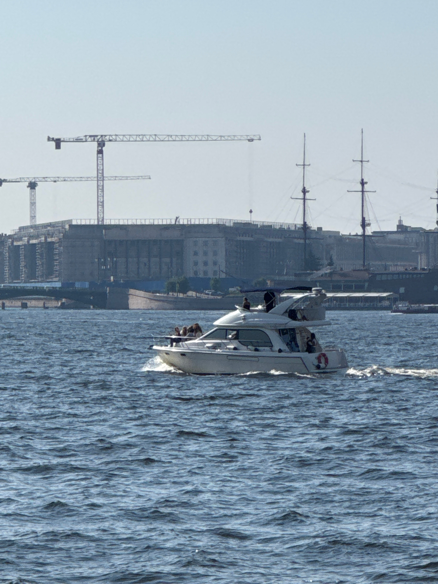 The image shows a cityscape with a body of water in the foreground. A white motorboat is moving through the water, creating a wake. There are several people visible on the boat. In the background, there is a large construction site with cranes, and further back, city buildings and what appear to be masts of old sailing ships. The sky is a pale, hazy blue, suggesting it is daytime, possibly overcast or early morning/late afternoon. The water appears choppy. The overall scene depicts a busy urban waterfront, possibly undergoing development, with recreational activity on the water. Given the mention of Saint Petersburg, Russia, it is likely the Neva River or a canal.
