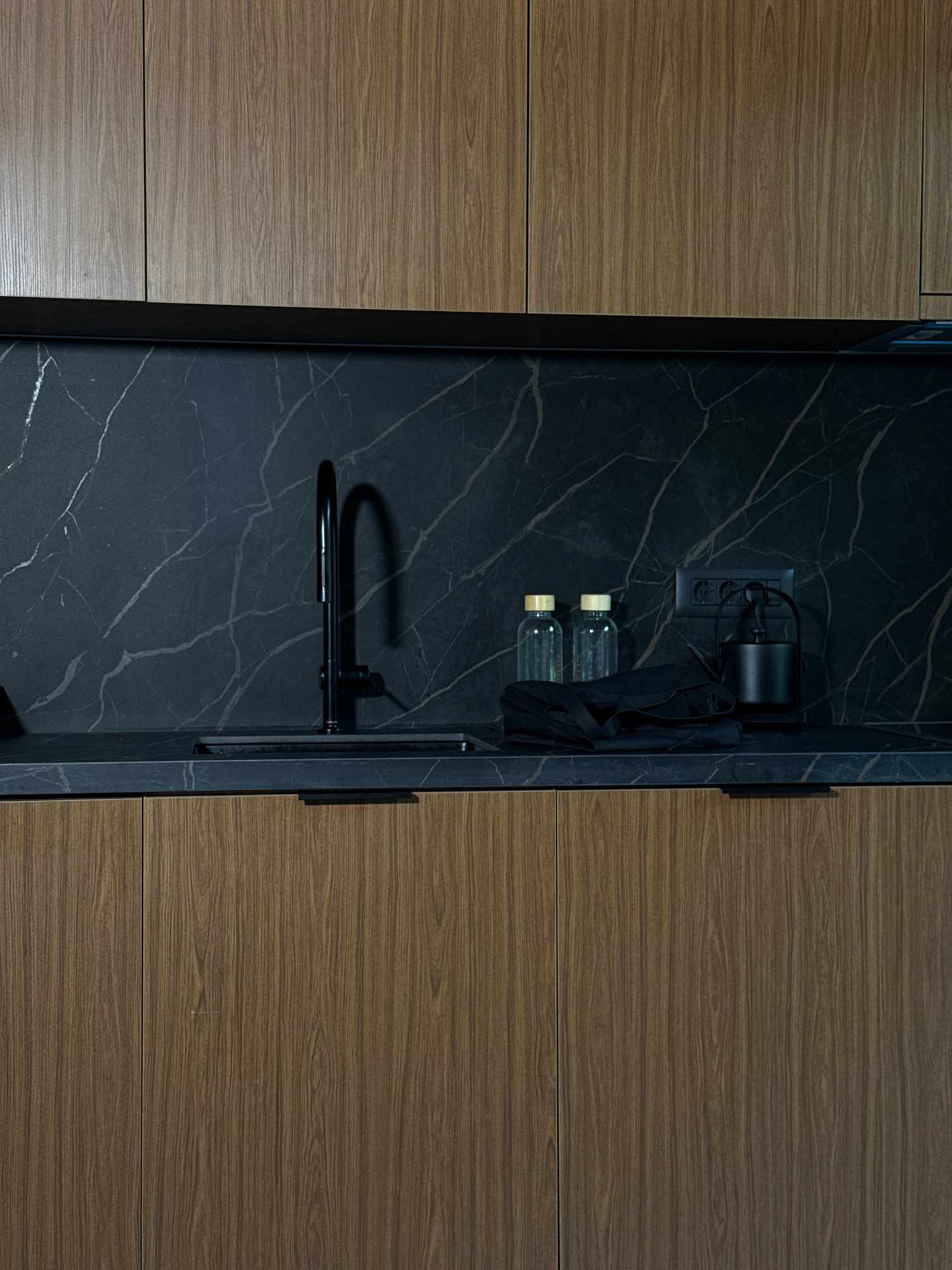 The image depicts a modern kitchen interior featuring dark marble countertops and backsplash, contrasted with wooden cabinetry. A black faucet stands over a sink. To the right, on the countertop, are two clear glass bottles with wooden stoppers, a folded black cloth, an electrical outlet with a black kettle plugged into it, and a black appliance. The scene is well-lit, highlighting the textures and finishes of the materials. The location is identified as Bakovići, Montenegro.