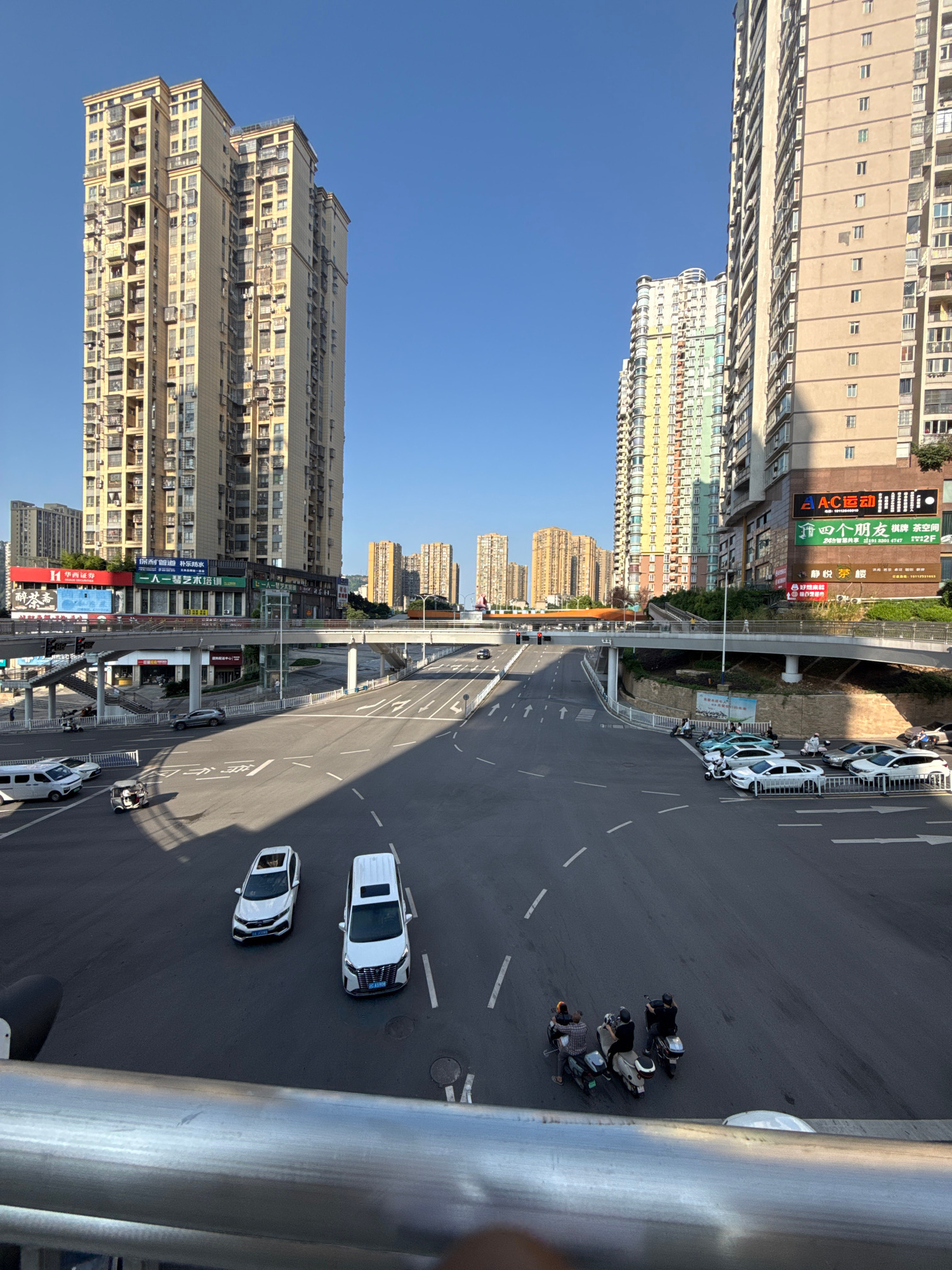 The image shows a daytime, slightly elevated view of a multi-lane intersection in Jiangjin, China, with several tall apartment buildings lining the background. The sky is clear and blue. In the foreground, a metal railing is visible. The intersection features road markings for multiple lanes and traffic flow. Several vehicles are present, including white vans, cars, scooters, and a three-wheeled vehicle. Some vehicles are parked along the side of the road, while others are in motion. On the left side of the intersection, there's a pedestrian overpass with stairs. The buildings in the background are modern, with numerous balconies and windows, and feature various advertisements and signs. One building on the right has a prominent sign with Chinese characters and a logo for "AC Sports."