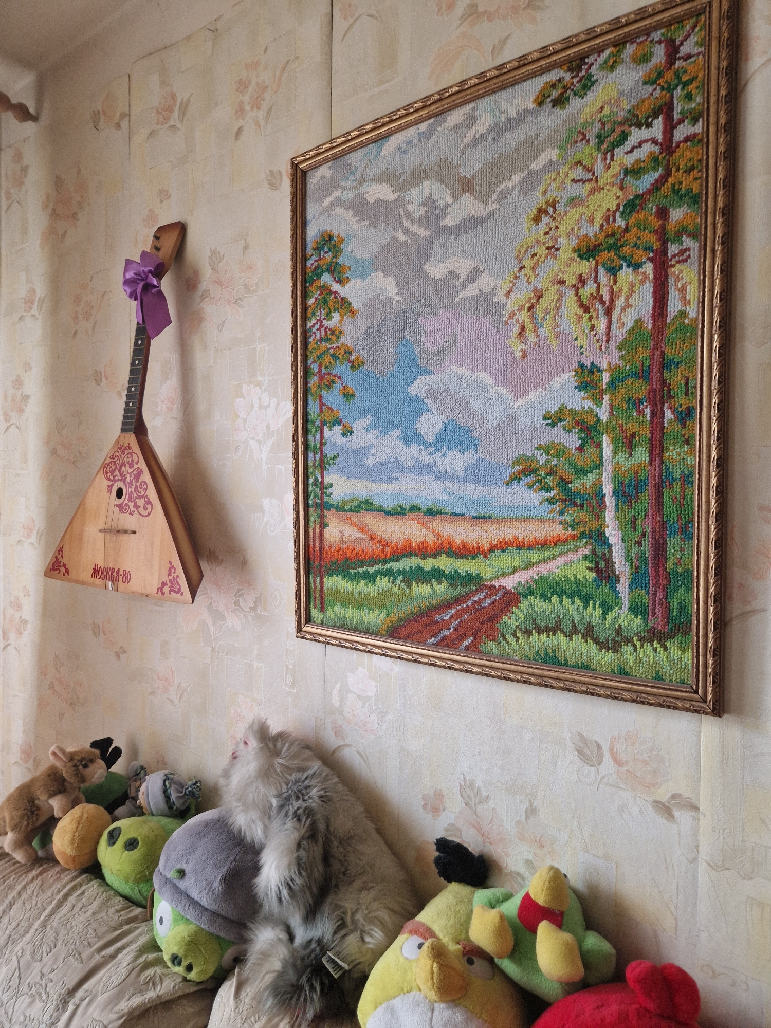 In an indoor setting, a wall adorned with floral patterned wallpaper serves as the backdrop for several objects. Mounted on the wall is a framed tapestry depicting a landscape with trees, a field, and a dirt path under a cloudy sky. To the left, a wooden balalaika instrument, decorated with a pinkish-purple bow and ornate pink designs, is hung on the wall. Below the balalaika and tapestry, a couch is covered with a textured fabric and is populated by a collection of stuffed toys. These include several green pig-shaped toys, a light brown animal plush toy, a fluffy grey and white cat plush toy, and various other colorful stuffed birds. The overall scene suggests a domestic interior, possibly a living room.