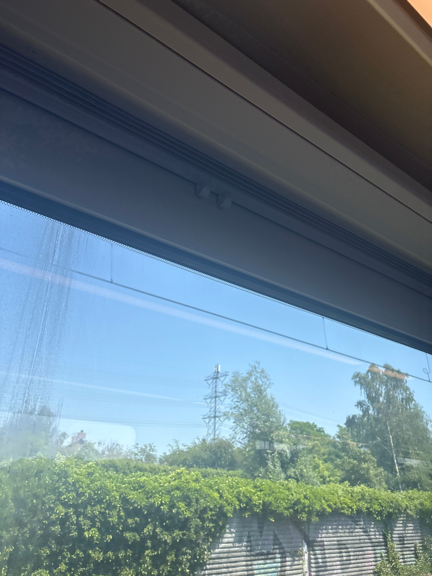 The image shows a view through a window, with the interior of a room on the left and an outdoor scene on the right. The interior features a grey, angled structure, possibly a window frame or part of a blind mechanism, with some white plastic clips visible. The outdoor scene is dominated by lush green foliage, including trees and bushes, suggesting a natural or well-maintained garden setting. A tall electrical pylon is visible in the distance, indicating an urban or suburban environment. The sky is a clear, bright blue, suggesting a sunny day. The reflection on the glass hints at the time of day being daytime, likely mid-morning or afternoon due to the clear sky and lighting. There are no people visible in the image. The overall mood is serene and bright, with the focus on the interplay between the indoor and outdoor environments. The photo appears to be taken in Eindhoven, Netherlands, consistent with the presence of electrical pylons often found in Dutch landscapes. No text is visible in the image.