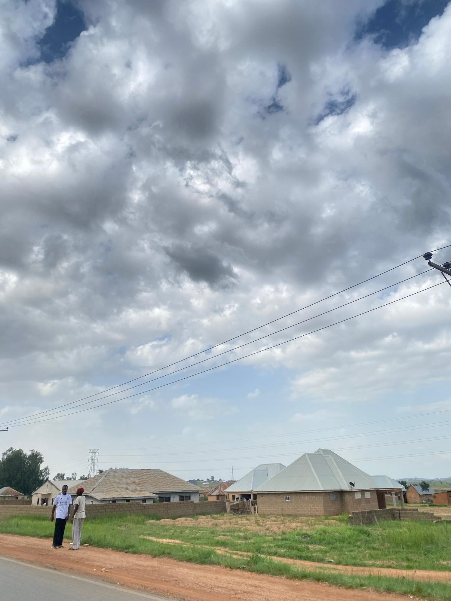 The image depicts a daytime scene in Rahwol Chom, Nigeria, featuring a cloudy sky with patches of blue. In the foreground, two individuals are standing on a dirt road next to a grassy area. They are dressed casually, one in a white t-shirt with a logo that appears to read "ADIDAS" and black pants, and the other in a light-colored outfit. Behind them, several buildings are visible, including structures with corrugated metal roofs and some under construction. Power lines stretch across the frame, and a distant electrical pylon can be seen. The overall impression is of a rural or semi-urban environment on an overcast day.