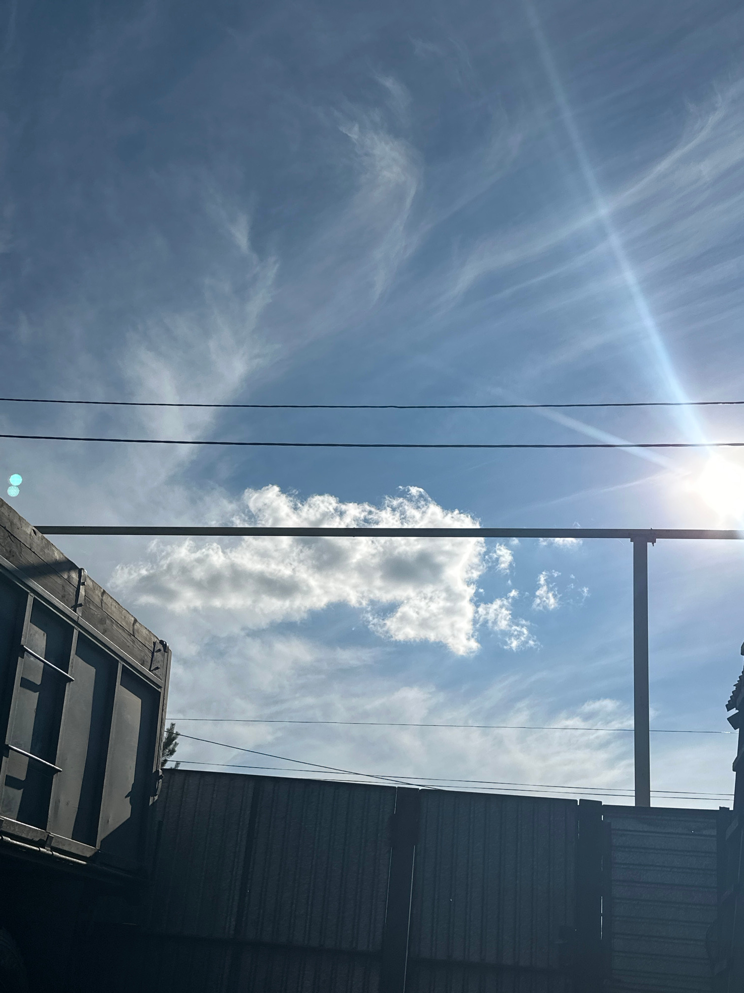 The image displays an outdoor scene featuring a bright blue sky with scattered white clouds. Wispy cirrus clouds are visible across the upper portion of the sky, and a prominent sunburst emanates from the upper right corner. In the foreground, a portion of a dark-colored industrial-looking structure, possibly a trailer or a shipping container, occupies the left side of the frame. A grey metal fence with vertical corrugations runs along the bottom of the image, and a slender vertical pole extends upwards through the center. Two parallel dark wires are suspended horizontally across the sky, with a thicker horizontal metal bar running below them. There are no people or animals present in the image. The environment suggests an industrial or utility area, consistent with a location in the city of Tobyl, Kazakhstan.