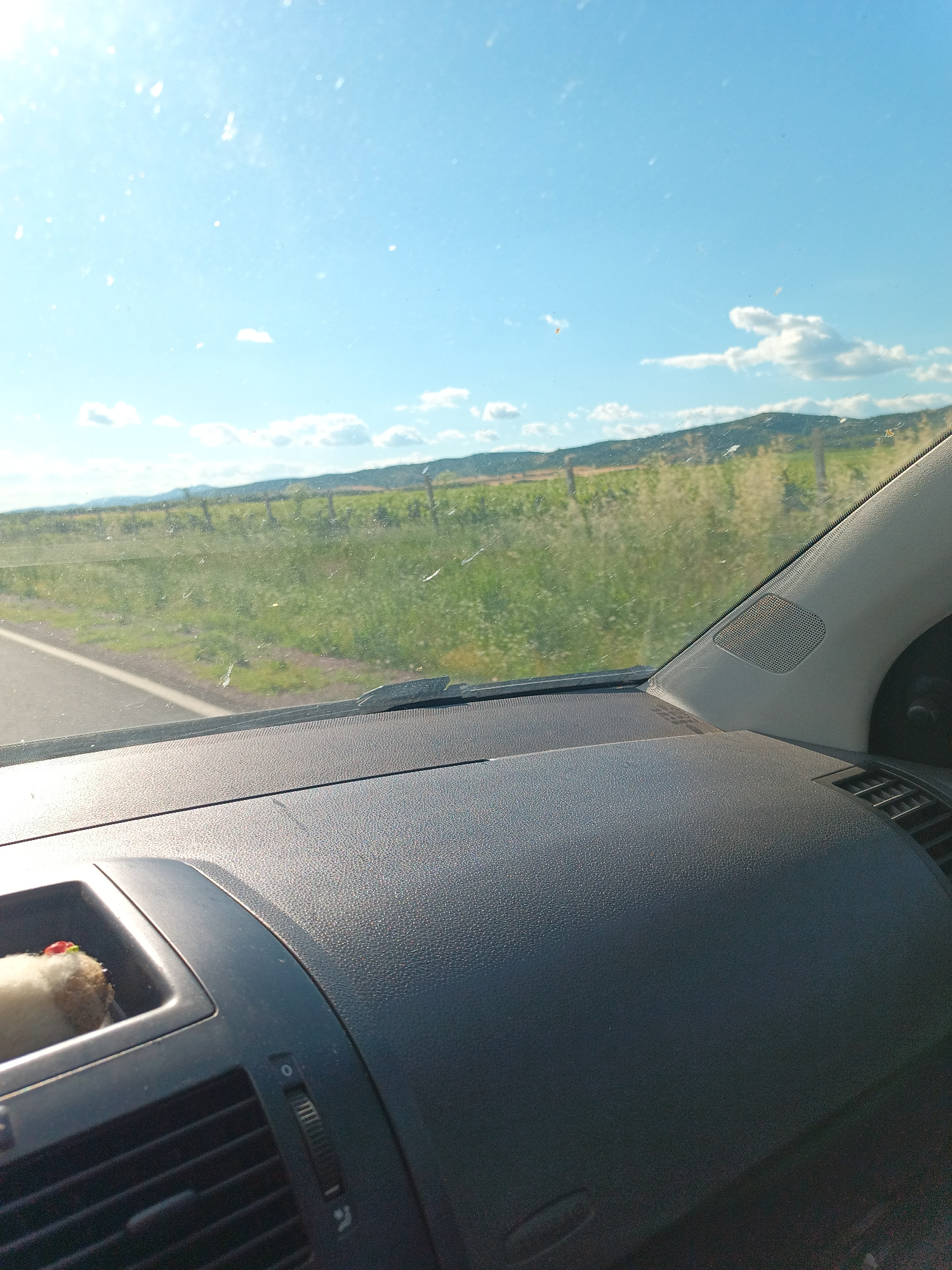The image captures a view from inside a car, looking out through the windshield. The scene is dominated by a bright blue sky with scattered white clouds, suggesting fair weather and likely daytime. A landscape of rolling hills covered in green vegetation stretches into the distance, with a fence line visible running horizontally across the middle ground. A road is visible on the left side of the frame, indicating the car is in motion.
Inside the car, the dashboard is in the foreground. A dark gray textured dashboard fills the bottom portion of the image. On the left, a small compartment on the dashboard holds a fuzzy object with a red tip. To the right of this compartment, a car air vent is visible. The overall impression is a snapshot of a scenic drive on a sunny day. The location cues, such as the type of vegetation and rolling hills, are consistent with rural settings in Bulgaria, and specifically Lozenets.