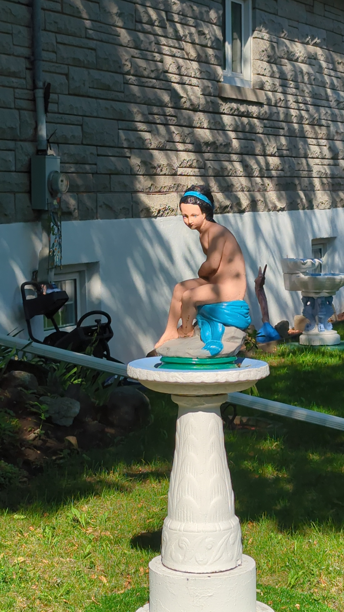 A statue of a young woman in a blue sash is perched on a rock on top of a white birdbath. The statue is in the yard of a house, with a stone wall and a window visible in the background. The yard is covered in grass and there are some bushes and rocks. The scene appears to be in Montreal, Canada, given the presence of a building and yard typical of residential areas in the city. The statue is the main subject, with the birdbath and house as secondary elements. There are no people present other than the statue.
