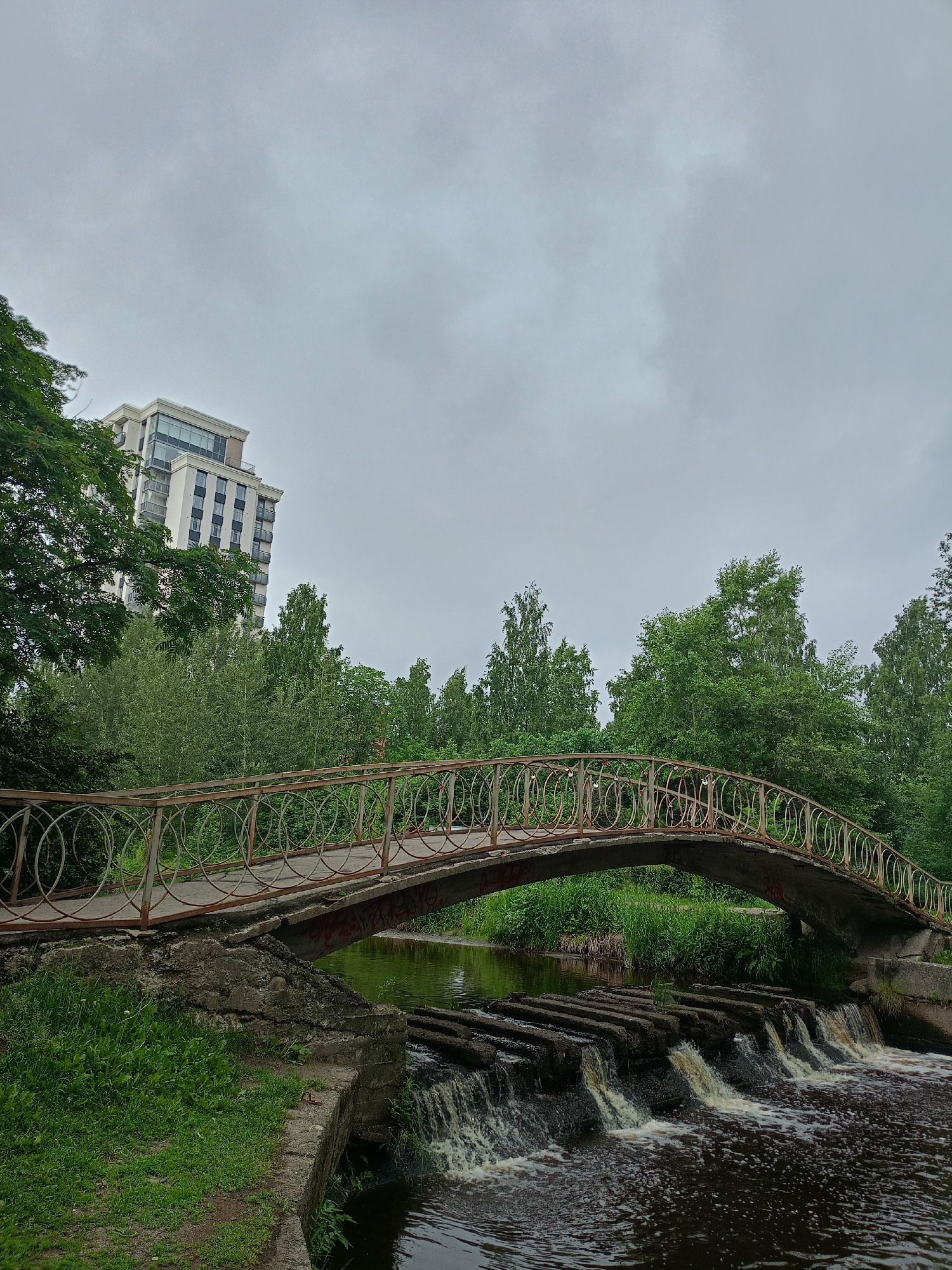 A concrete pedestrian bridge arches over a dark river with a small, segmented waterfall. The bridge has a rusty metal railing with circular decorative elements. To the left of the bridge, lush green grass and trees line the bank, and a tall, modern building with many windows stands behind them. The background is filled with dense green trees, and the sky is overcast and gray. The scene appears to be a park or natural area adjacent to an urban development. No people are visible, and the overall mood is calm and perhaps a little somber due to the cloudy weather. There is some red graffiti on the underside of the bridge. The time of day is indeterminate due to the overcast sky.