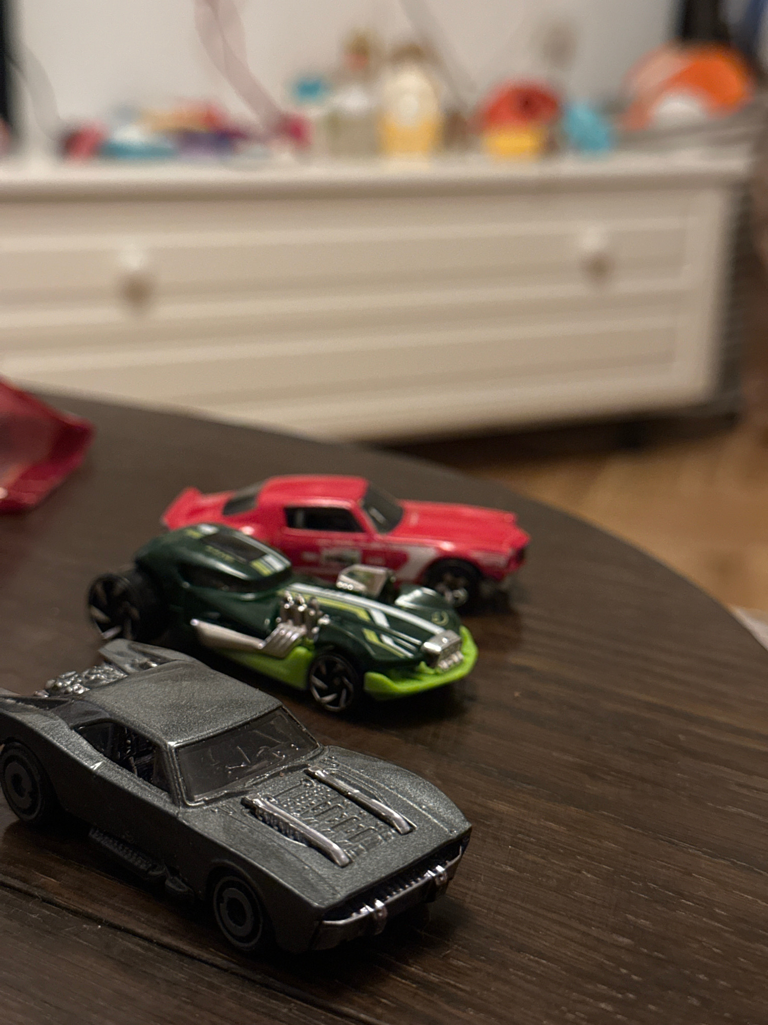 A close-up, eye-level shot shows three toy cars on a dark wooden surface. The cars are arranged in a line from closest to farthest. The closest car is a metallic gray Batmobile, with visible engine details on the hood and a prominent spoiler at the rear. Behind it is a dark green and lime green race car with chrome accents. The farthest car is red with white racing stripes. The background is blurred, showing a white dresser with drawers and some colorful objects on top. The scene is indoors, with no discernible view of the city of Sofia, Bulgaria.