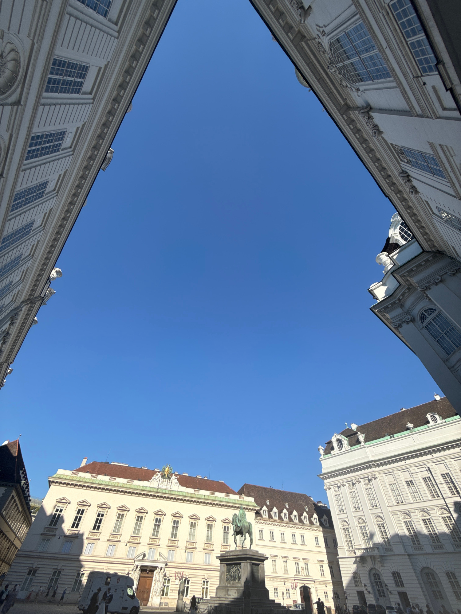 This image shows a low-angle view looking up at classical architecture against a clear blue sky. The scene is framed by buildings on all sides, suggesting a courtyard or narrow street. The architecture features ornate facades, multiple windows with detailed mullions, and decorative cornices. On the right side of the image, a statue of a person on horseback is visible, along with elements of another building that appears to be a monument or a significant structure. A white van is parked in front of one of the buildings.
The time of day appears to be late morning or early afternoon, as indicated by the strong shadows cast by the buildings. The weather is fair with a cloudless sky. The location is suggested to be Vienna, Austria, based on the architectural style commonly found in European capitals. No text is visible in the image. The overall impression is one of historical grandeur and urban density.