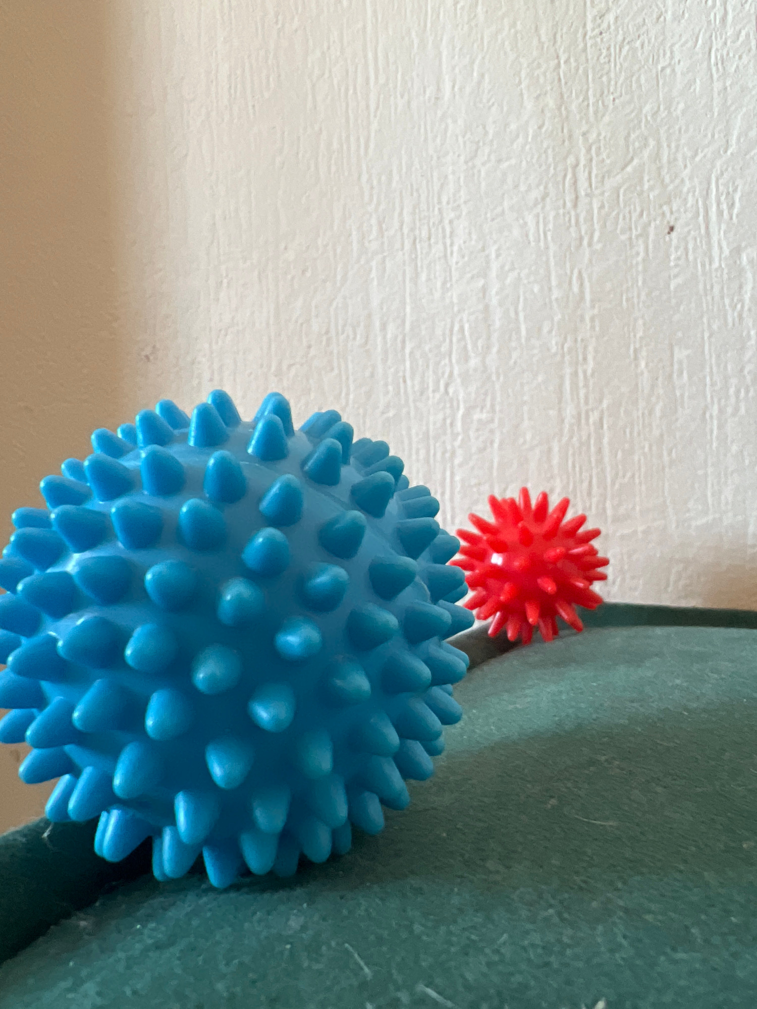 The image displays two spiky massage balls, one large and blue, positioned in the foreground, and a smaller red one in the background. They are resting on a textured, dark green surface, possibly fabric or upholstery. Behind the balls, a light-colored wall with a visible textured pattern extends upwards. The lighting appears to be natural, casting subtle shadows. The scene is devoid of any human presence or activity. There is no identifiable information within the image to suggest a specific location within the city of Novosibirsk, Russia.