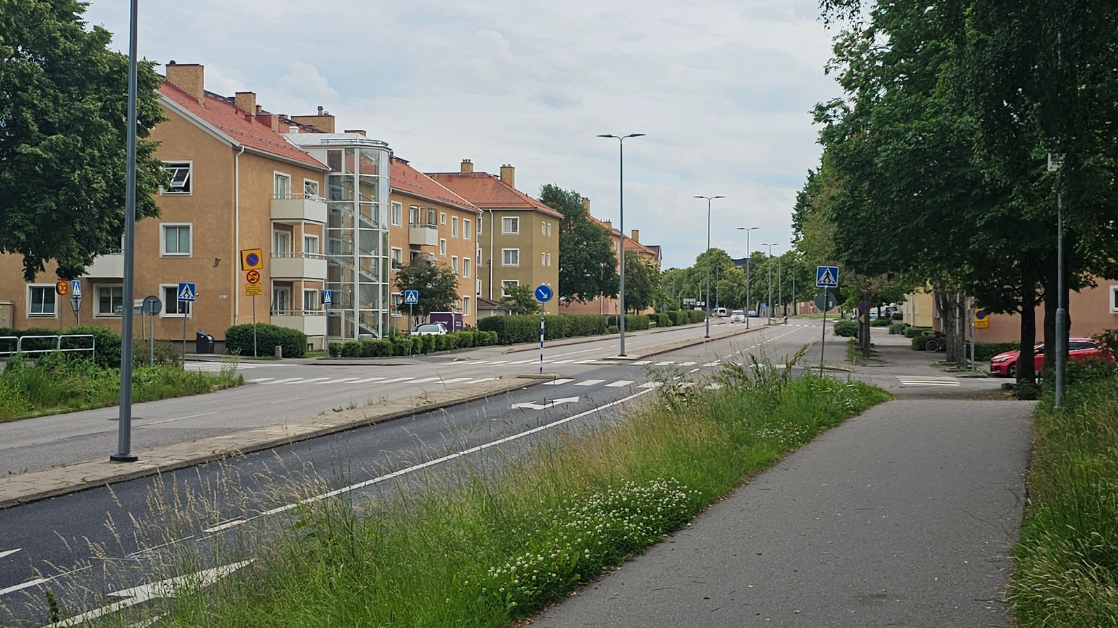This media file captures a street scene in Linköping, Sweden, on a cloudy day. The image depicts a residential street lined with multi-story apartment buildings, characterized by their yellow facades and red tiled roofs. An external glass elevator adds a modern architectural touch to one of the buildings.
The street features a pedestrian crossing, traffic lights, and lampposts. The road is paved, with white lane markings. On the right side of the road, there's a sidewalk and a path for pedestrians and cyclists, bordered by a strip of green grass and trees. The grass along the sidewalk is dotted with small white wildflowers. On the left side of the road, more grass and trees are visible, along with a row of residential buildings.
Several road signs are present, including a "30" speed limit sign and pedestrian crossing signs. A blue and white striped pole with a directional arrow is also visible. Red cars are parked along the street, and a bicycle is leaning against a building.
The overall atmosphere is calm and ordinary, suggesting a typical day in a residential area. There are no people clearly visible in the frame, giving a sense of quietude. The overcast sky indicates a possibly cool or mild weather condition. The presence of trees and well-maintained sidewalks suggests a well-developed urban environment.