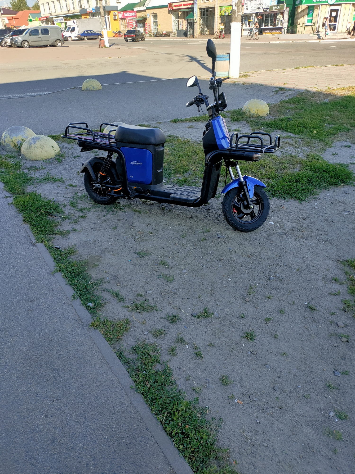 A blue electric scooter with black racks on the front and rear is parked on a dirt patch next to a paved walkway and a road in Nosivka, Ukraine. The scooter has a visible "CORBER R-3" logo on its side. In the background, buildings line a street with cars and pedestrians visible. Several rounded concrete bollards are scattered around the area, some appearing yellowed or weathered. The scene appears to be a public area with commercial establishments.