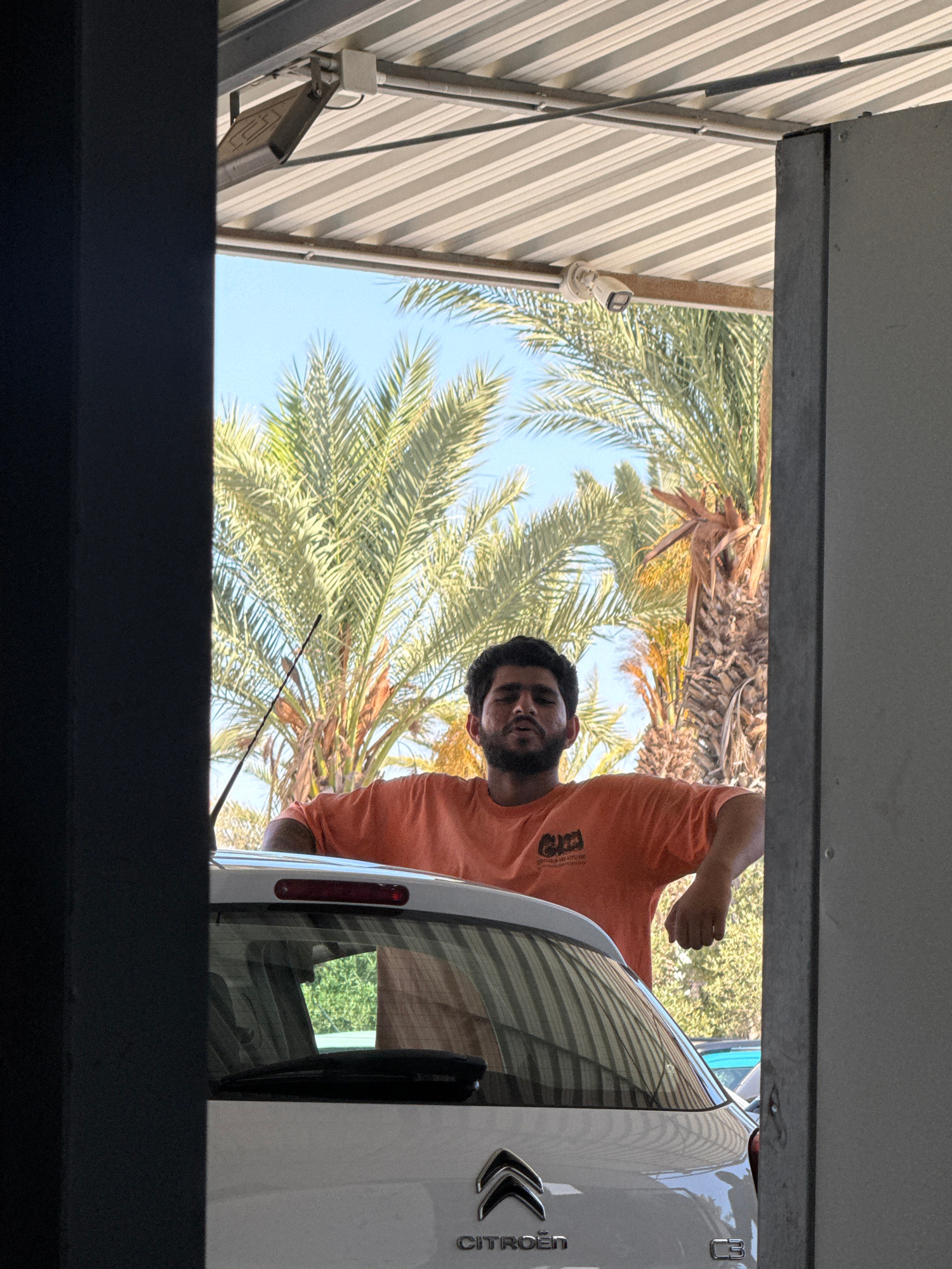 The image depicts a medium shot of a young man standing behind the rear of a white Citroen C3 car. He is wearing an orange t-shirt with a graphic logo. The man's arms are resting on the car's roof. His body is angled to his left, and he is looking down and to his right. The car's rear windshield and wiper are visible, along with the Citroen logo and "C3" model designation. The scene appears to be outdoors under a corrugated metal roof structure, possibly a carport or part of a building. Behind the man and car, palm trees and a clear blue sky are visible, suggesting a warm climate. The framing of the shot, with dark vertical elements on the left and right, implies the viewer is looking through an opening or doorway. The location is identified as Timi Community, Cyprus.