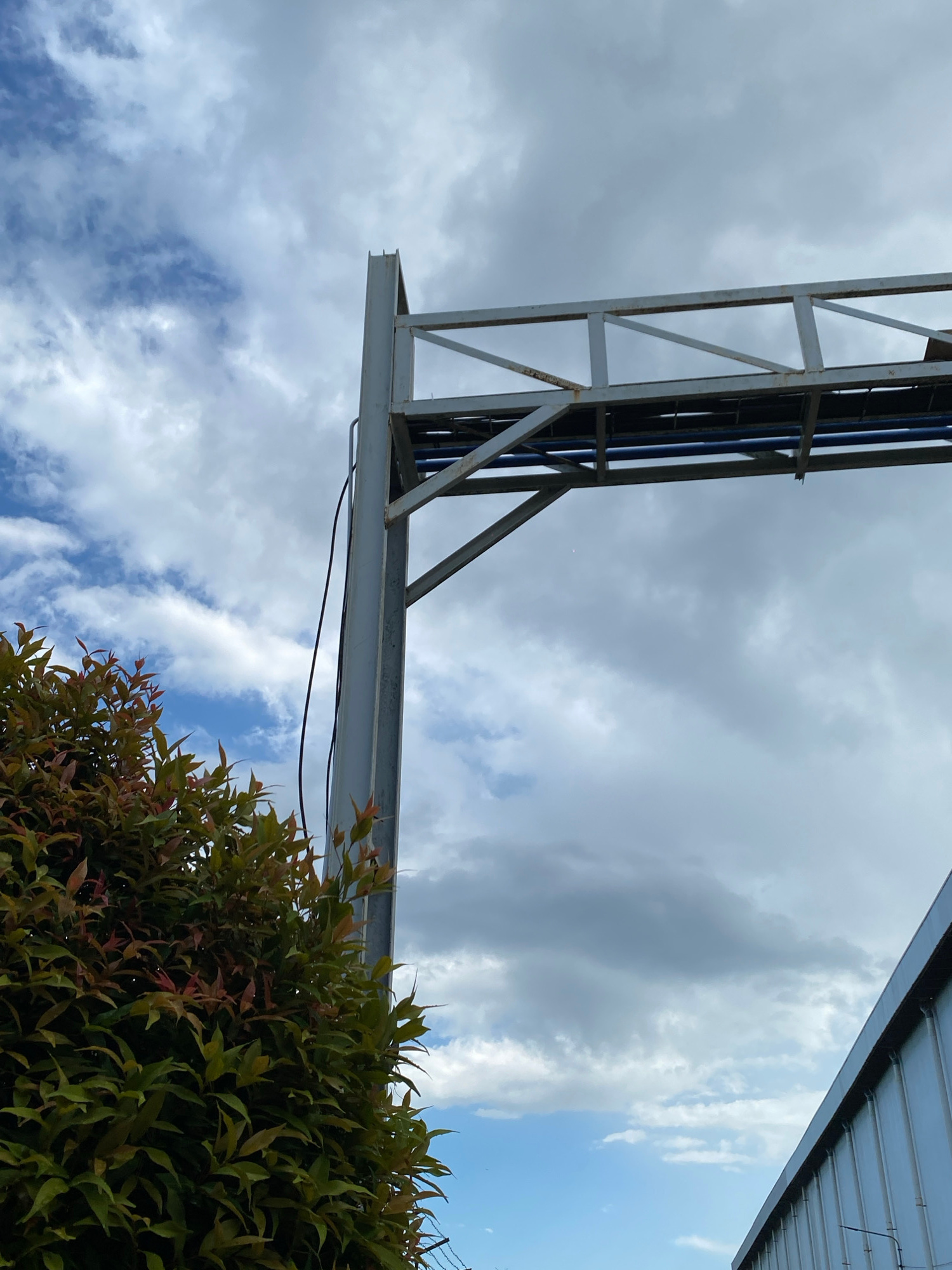 The image captures an outdoor scene featuring a metal overhead structure, likely a sign or conduit support, against a cloudy sky. To the left, a dense green bush with reddish new growth is partially visible. A gray metal pole supports the overhead structure, with a black cable running down its side. The overhead structure itself is made of interconnected metal beams, with blue pipes visible underneath. In the lower right corner, the angled edge of a pale blue or silver building with vertical ribbed siding is present. The sky is a mixture of white and gray clouds with patches of blue sky. The image is taken from a low angle, looking upwards.
