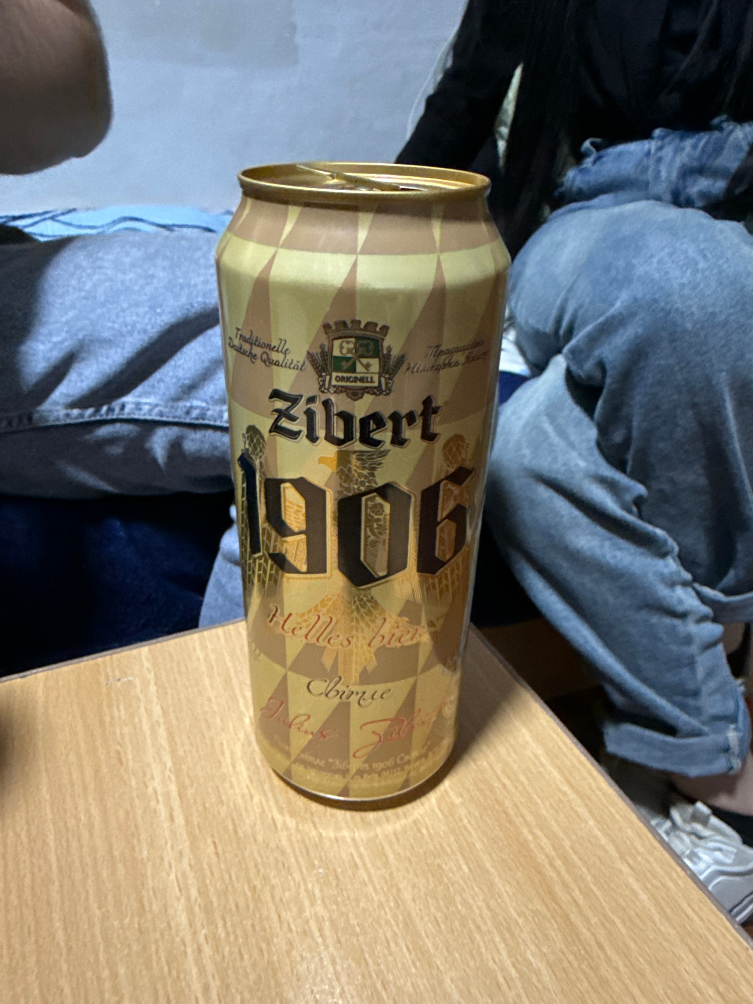A close-up, eye-level, medium shot captures a golden and brown patterned beer can positioned on a light-colored wooden table. The can is upright, with its top slightly ajar. Text on the can, in dark brown lettering, includes "Zibert 1906" and "Helles bier." The background is blurred, showing parts of people wearing denim and grey clothing, suggesting a casual indoor setting. The scene does not provide any visual cues or context to indicate a location in the city of Sambir, Ukraine.