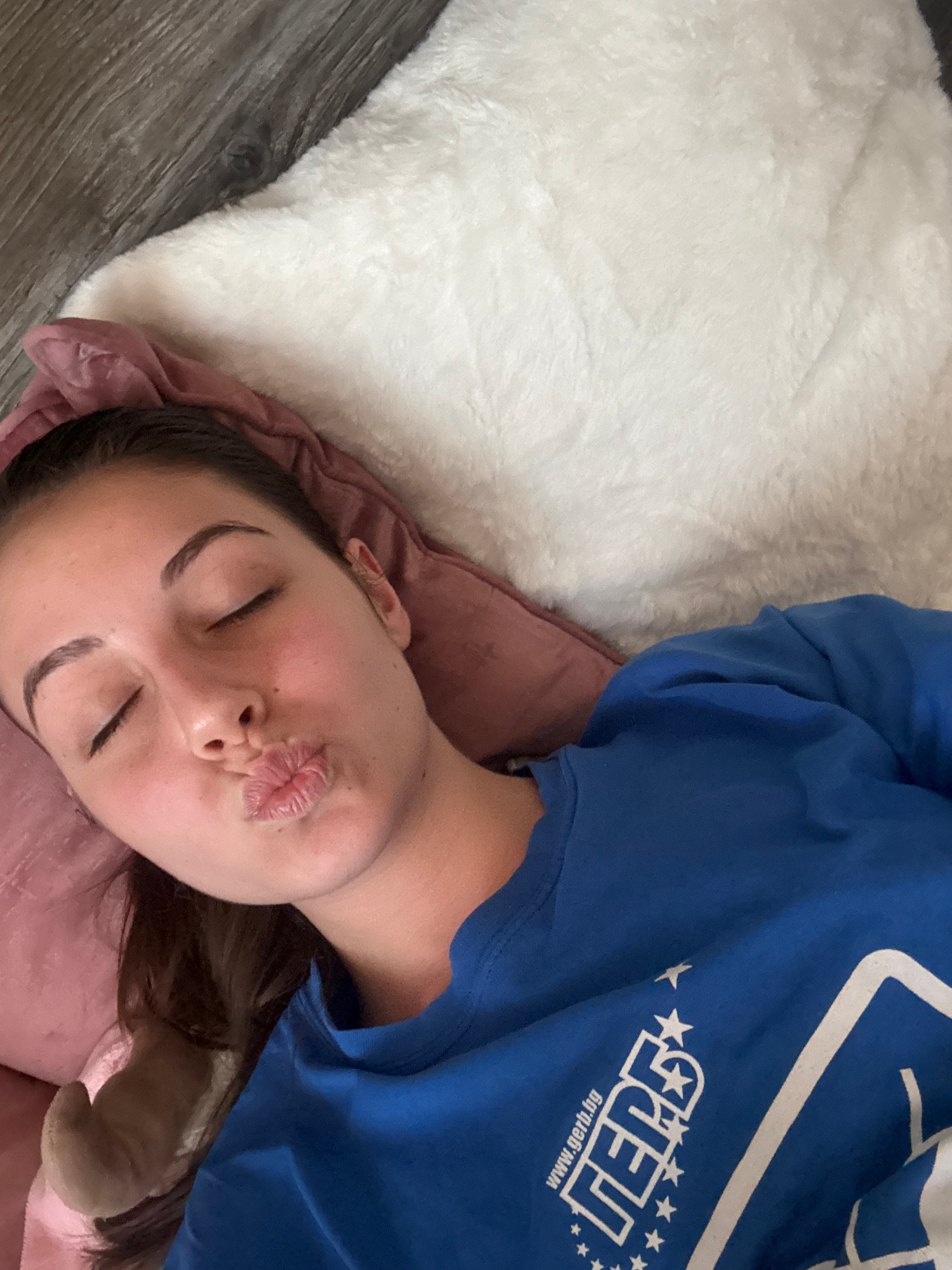 An overhead shot captures a young woman with her eyes closed and lips puckered in a kiss pose, lying on a soft surface that appears to be a bed or couch covered with a white fluffy blanket and a pink pillow. She is wearing a royal blue t-shirt with a white logo on the left sleeve. The logo features text that reads "GERB" with "www.gerb.bg" above it, surrounded by stars. The scene is indoors, likely in a bedroom, given the bedding. The floorboards visible in the upper left corner suggest a wooden floor. The overall impression is one of a casual, relaxed moment captured in a domestic setting.