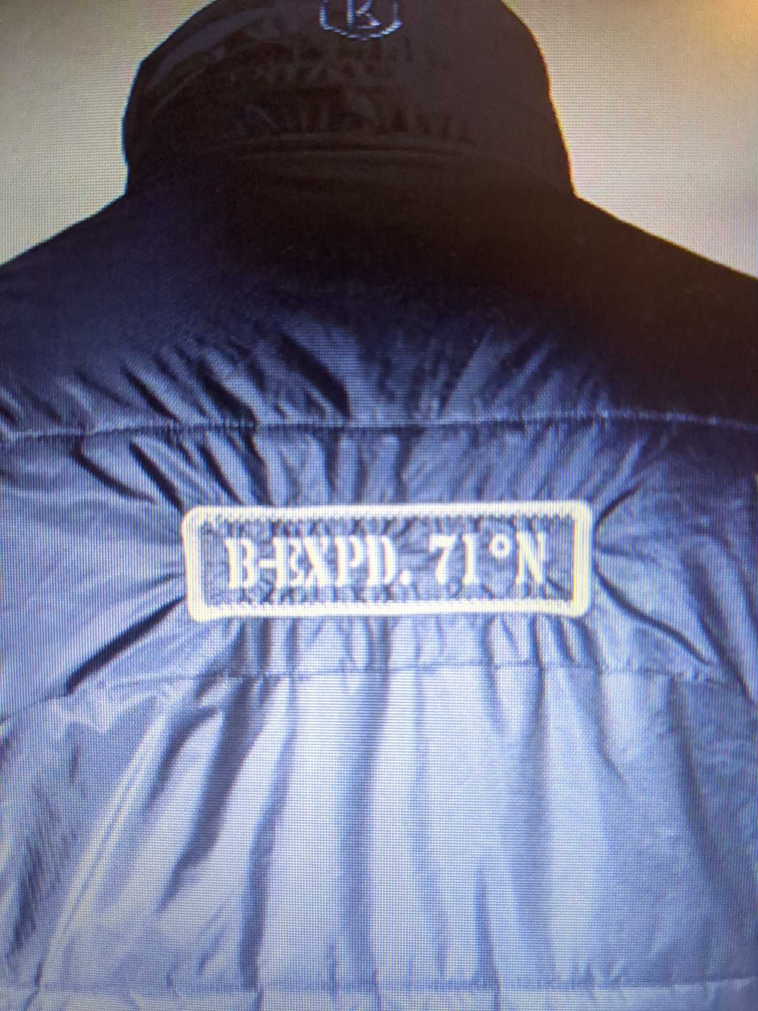 The image shows the back of a dark blue jacket with a rectangular patch on it. The patch has an embroidered design in a lighter color, possibly gold or yellow. The text on the patch reads "B-EXPD. 71°N". The jacket appears to be made of a shiny, somewhat crinkled material. The background is a muted, textured surface. There is a logo visible at the top of the jacket collar, which appears to be a stylized "R" within a circular shape. The image quality is low, with visible pixelation. The context of this image in Minsk, Belarus is not provided in the visual information.